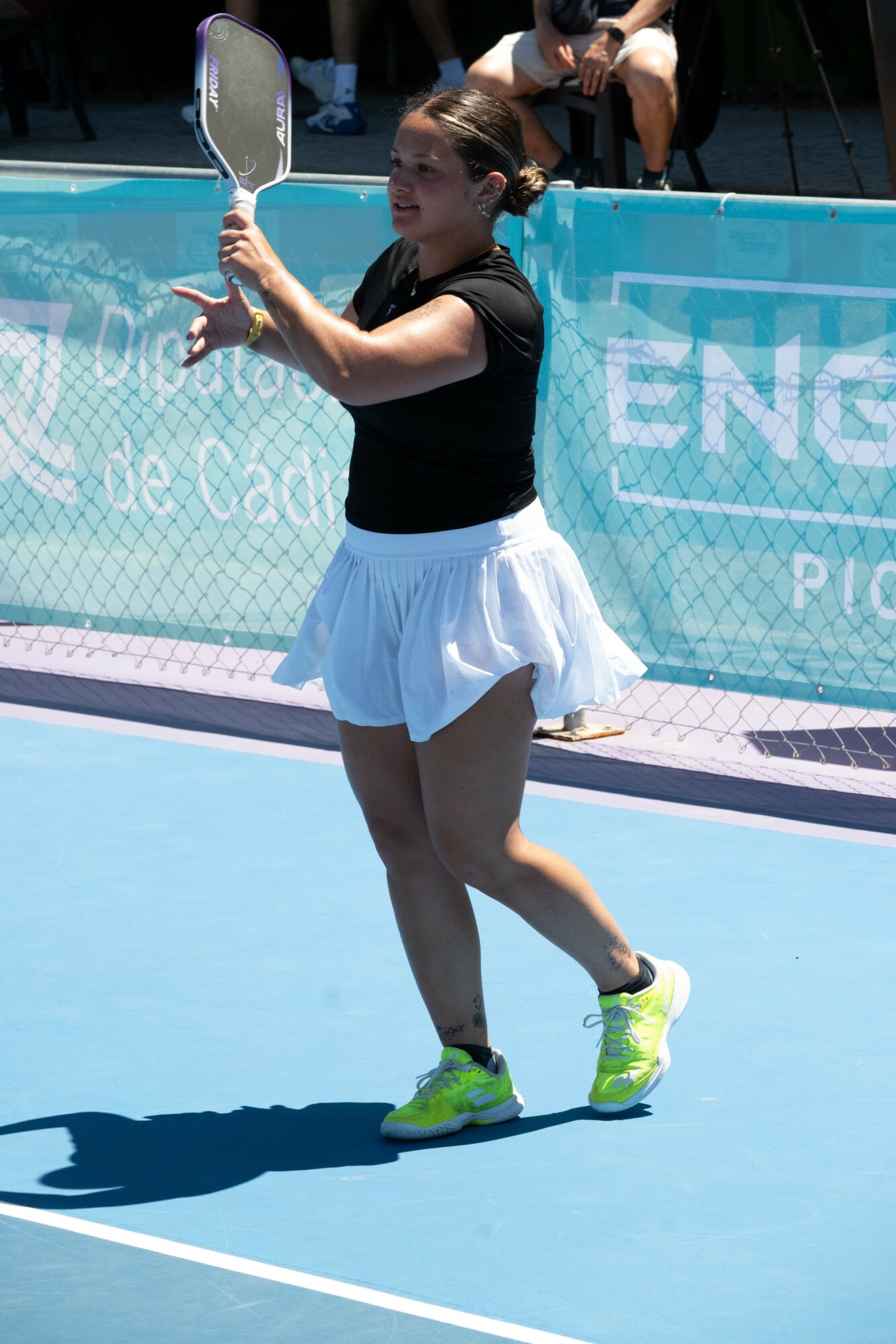 Female tennis player on court holding racket, wearing black top, white skirt, and yellow shoes, with a blue background.