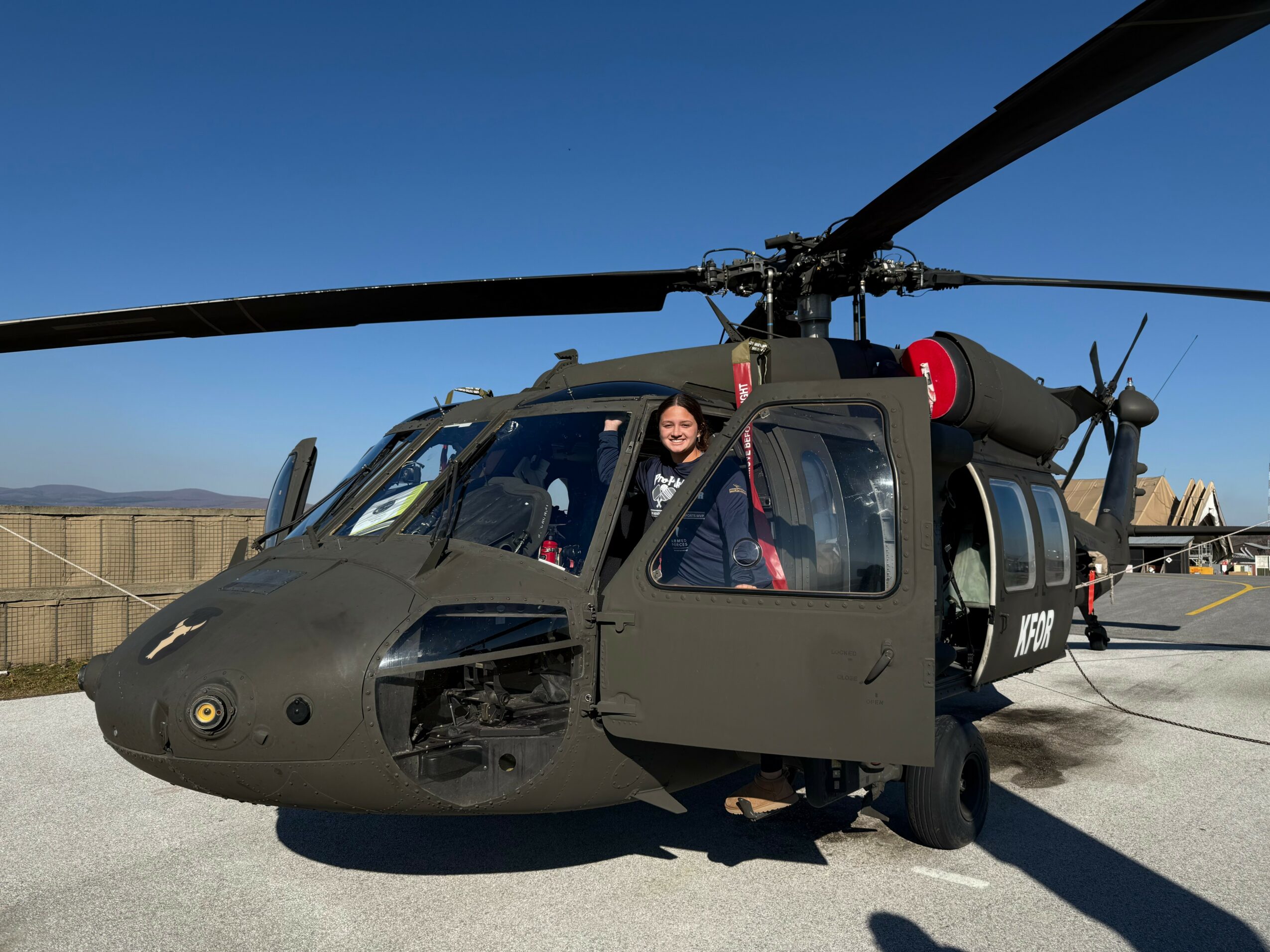 Person standing outside a military helicopter on a tarmac, with a fence and mountains in the background.