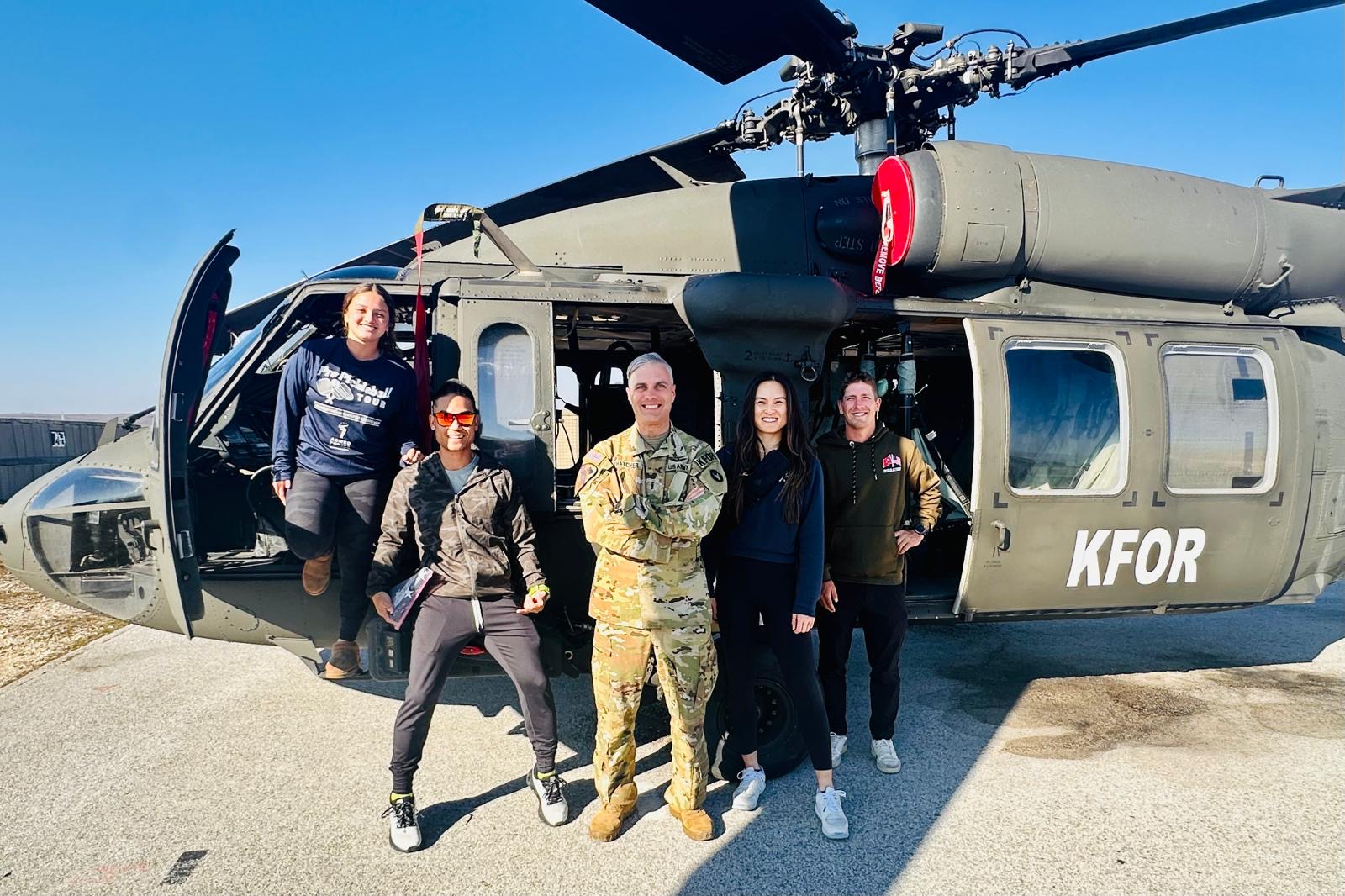 Group of five people standing in front of a military helicopter on a clear day.