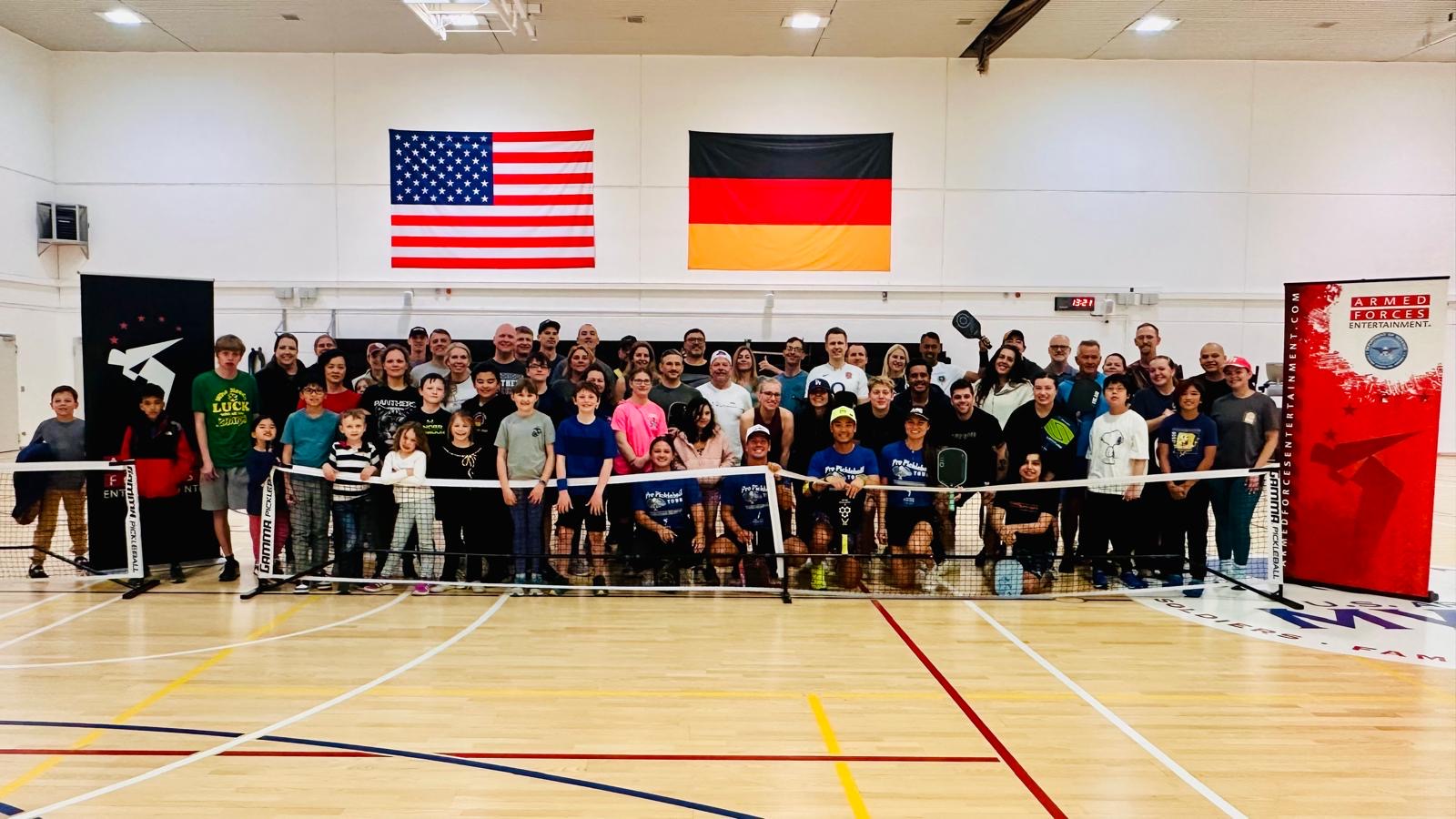 Group of people in a gymnasium with American and German flags hanging behind them, some holding a banner, children and adults present.