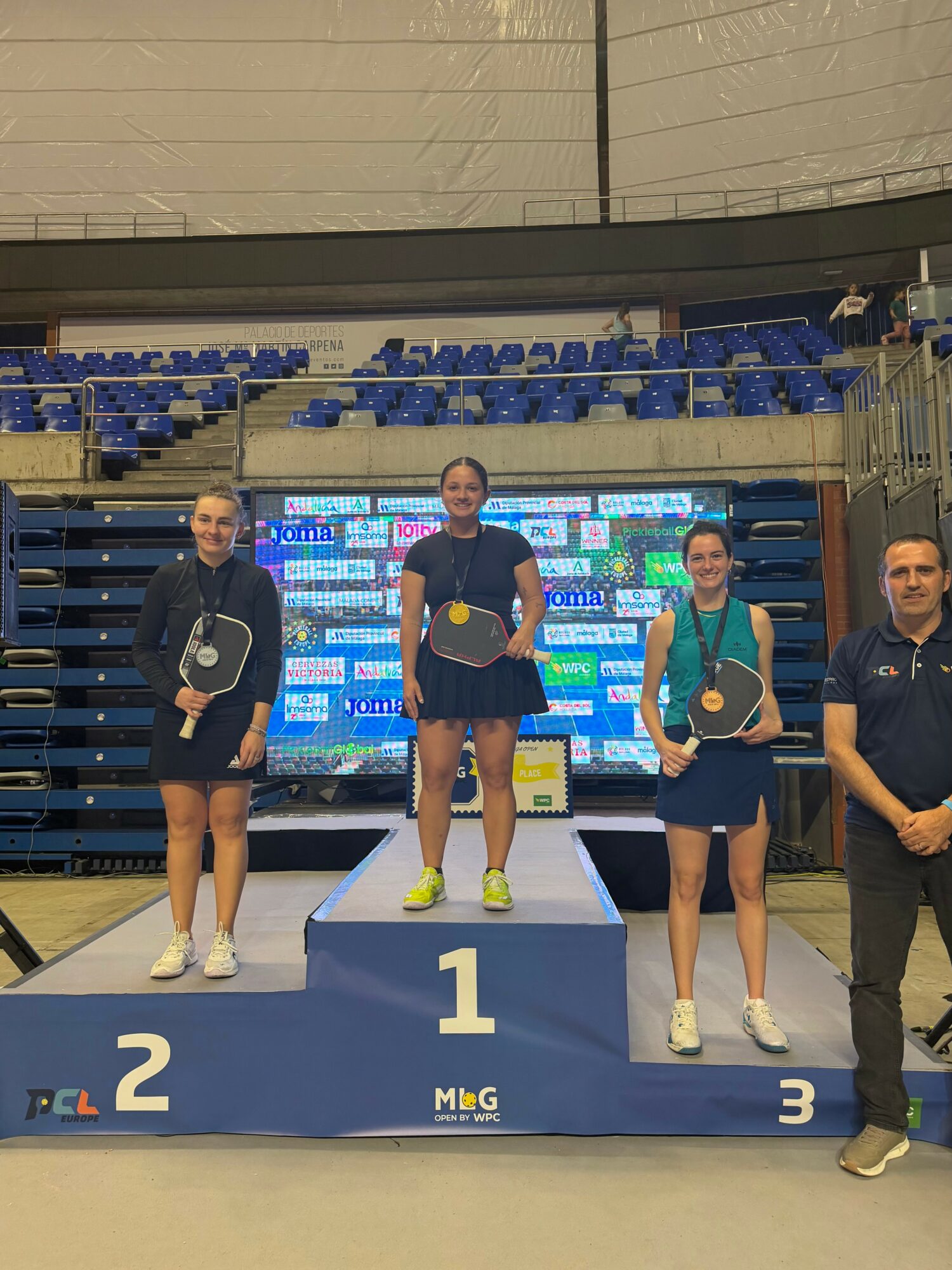 Three female athletes on a winners' podium holding rackets, with a man standing beside them, in an indoor sports arena.