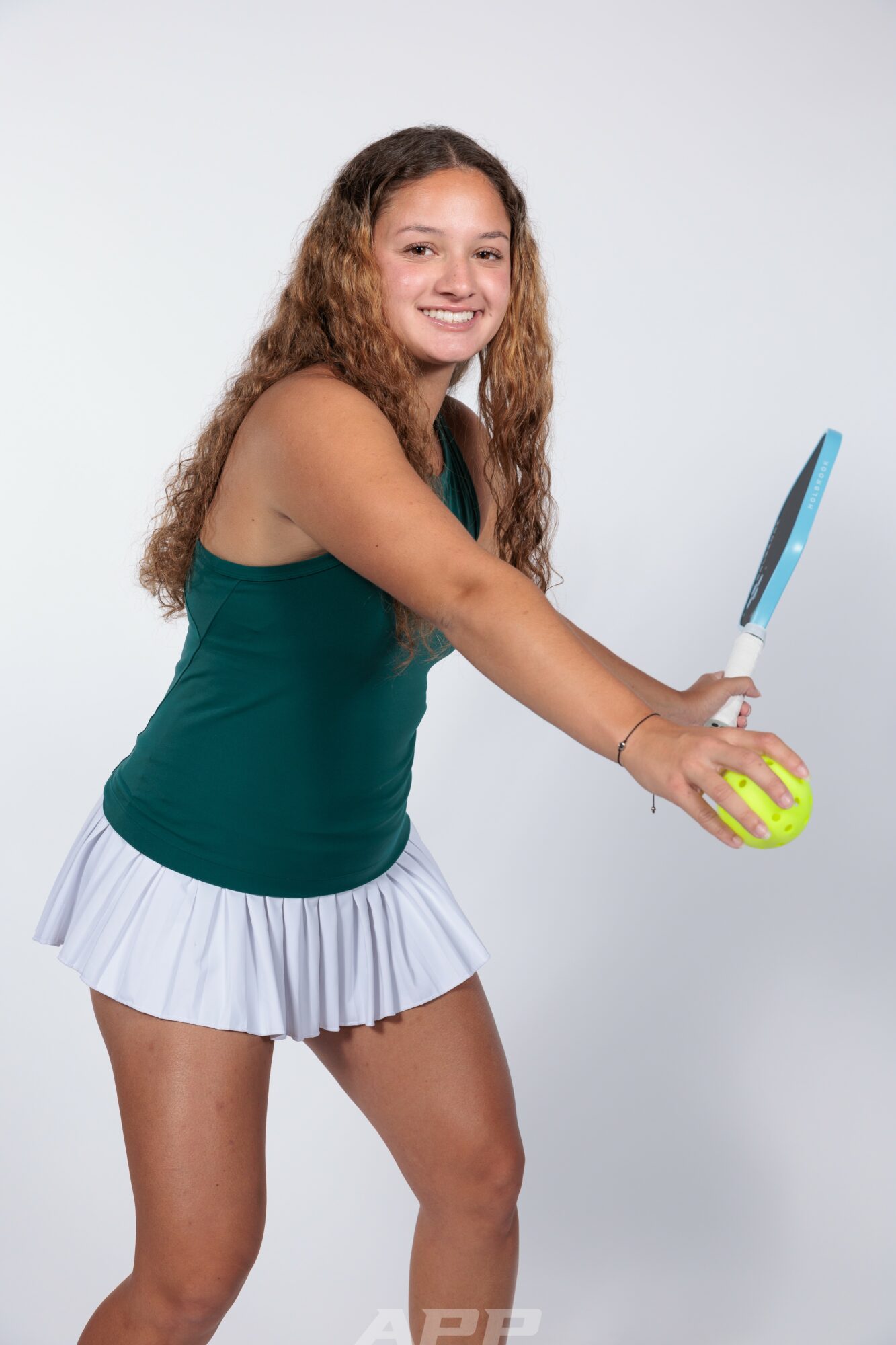 Young girl with long curly hair holding a tennis racket and tennis ball, smiling, wearing a green top and white skirt.