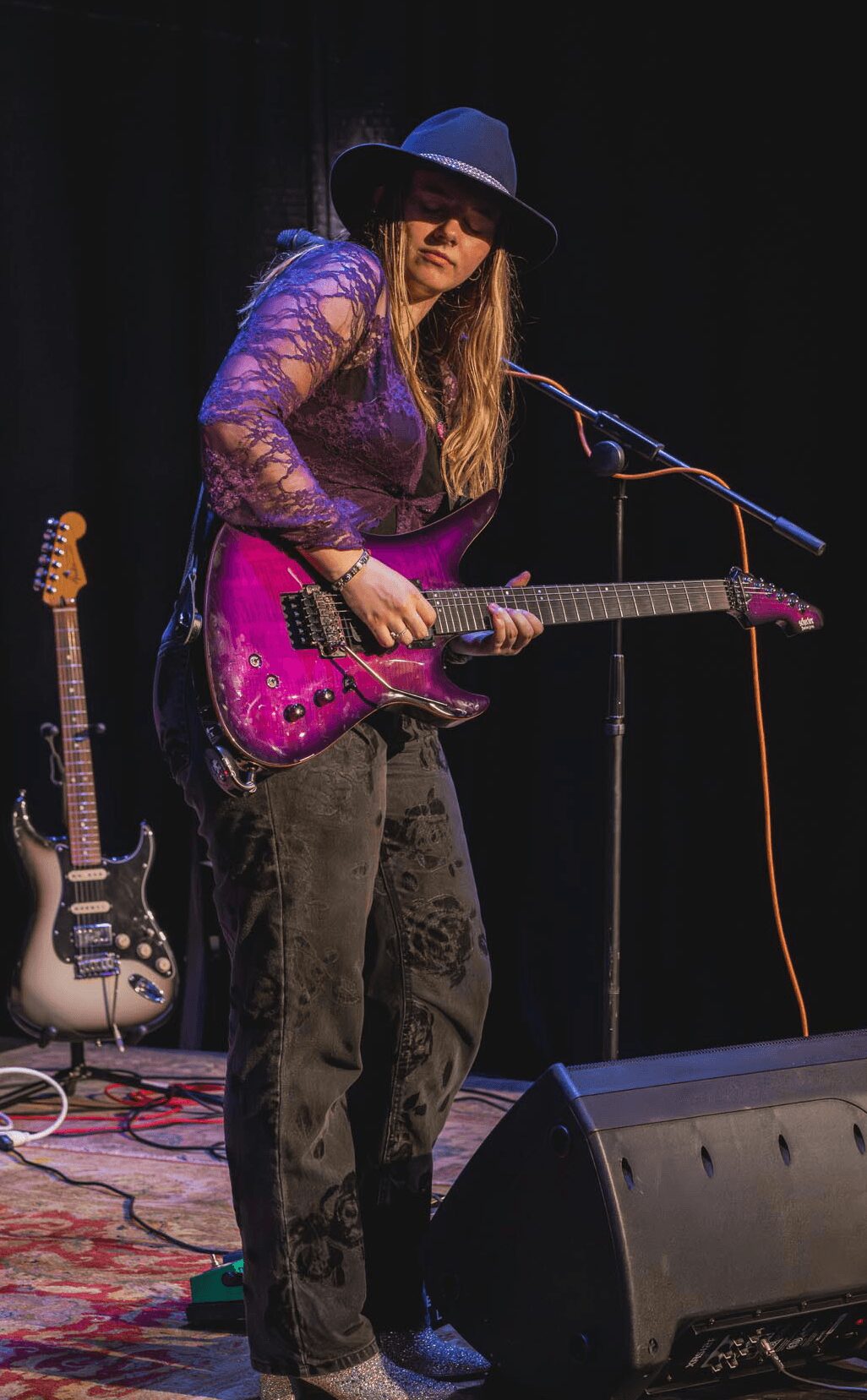 Female musician playing a pink electric guitar on stage, wearing a wide-brimmed hat and lace top, with a microphone nearby.