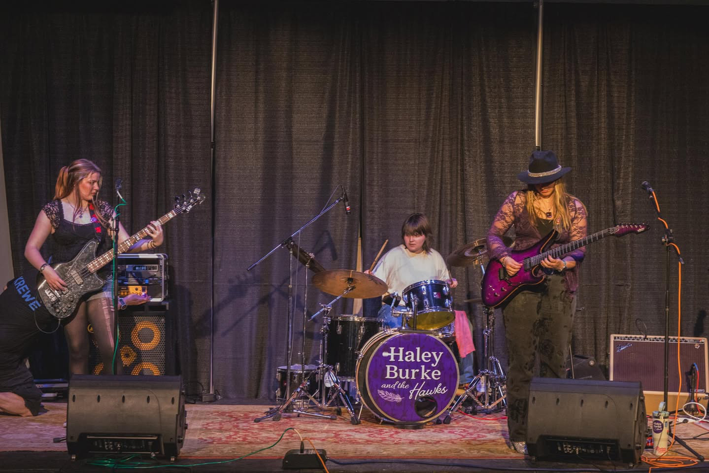 Three musicians perform on stage with instruments, a drum set, and a dark curtain backdrop.