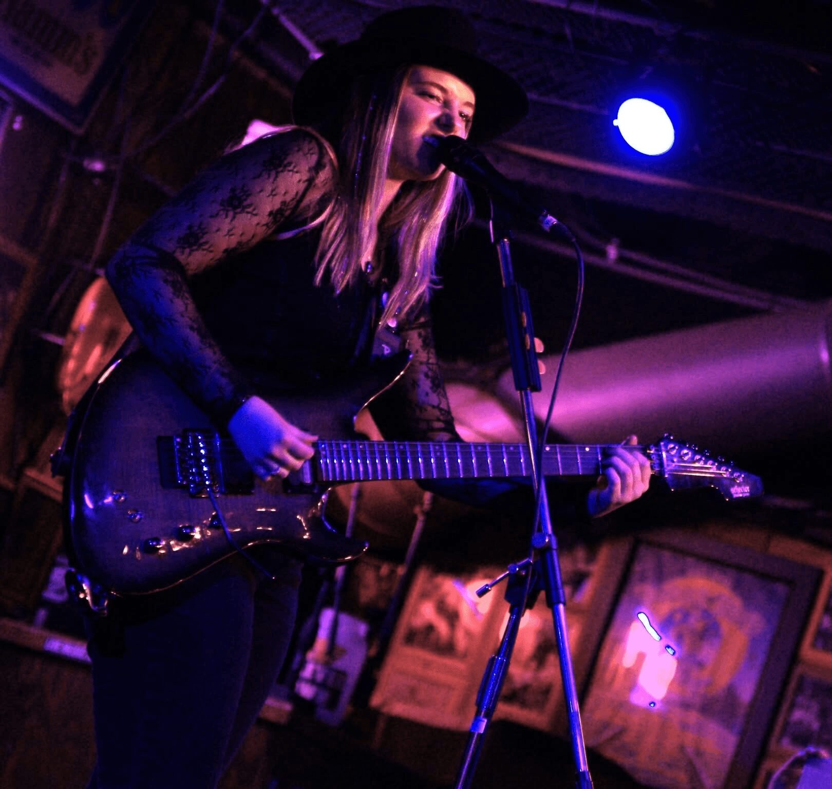 Woman singing and playing guitar on stage with purple lighting, wearing a hat and lace top.