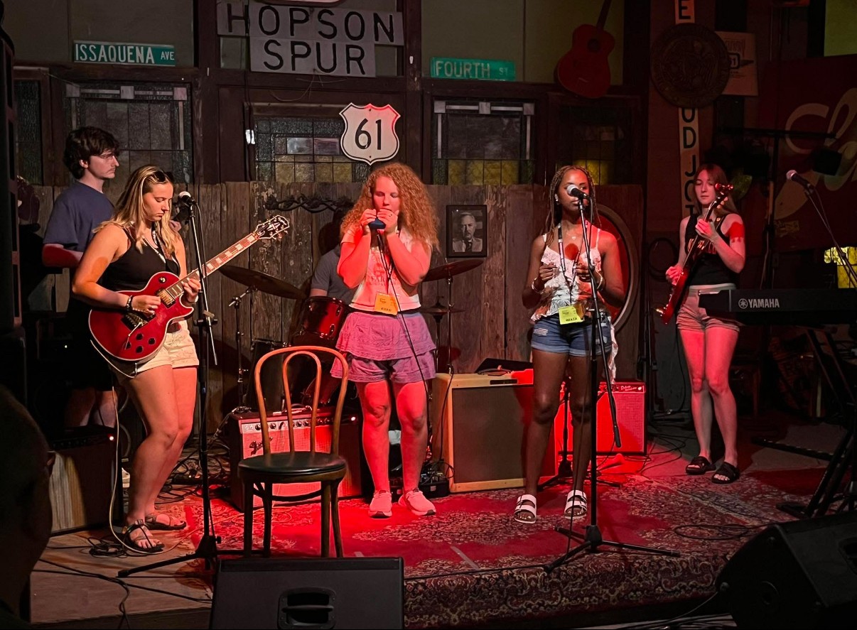 Group of five women performing on stage with musical instruments and microphones, in a cozy venue with signs and decorations.