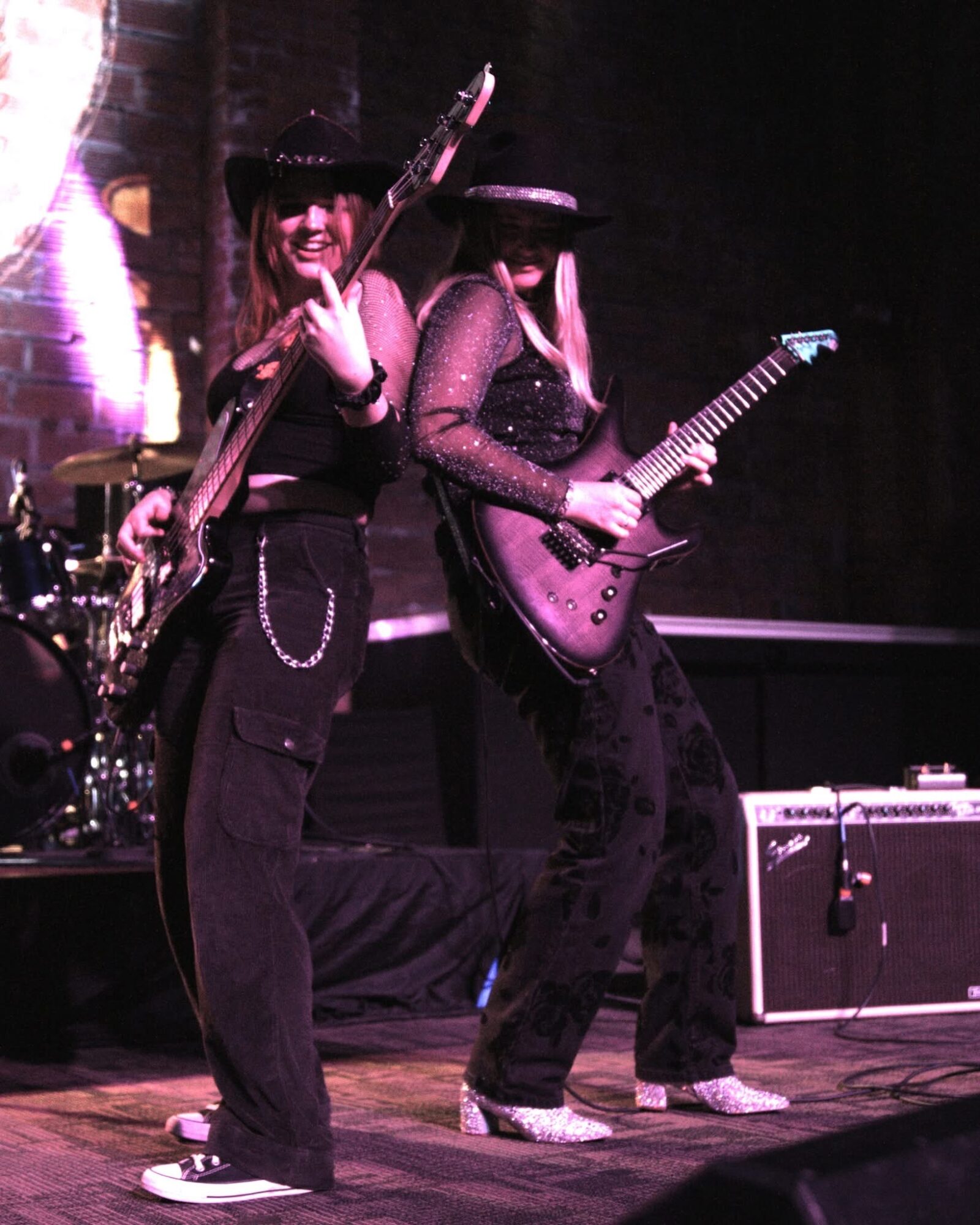 Two women playing electric guitars on stage, one wearing a hat and the other with long hair, with a drum set in the background.
