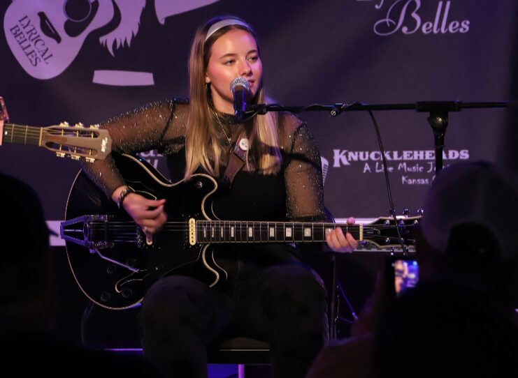 Young woman playing guitar and singing into microphone on stage with purple background and logos