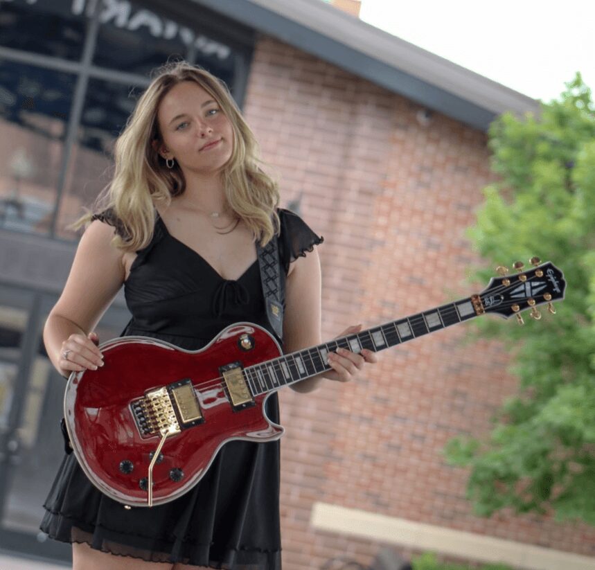 Young woman with wavy blonde hair holding a red electric guitar outdoors, brick building and green trees in background.