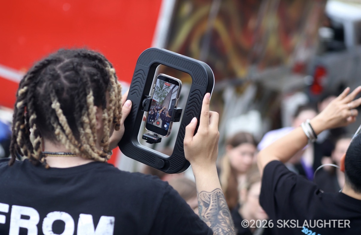 Person with braided hair holding a camera, taking a photo at an outdoor event with a crowd and colorful background.