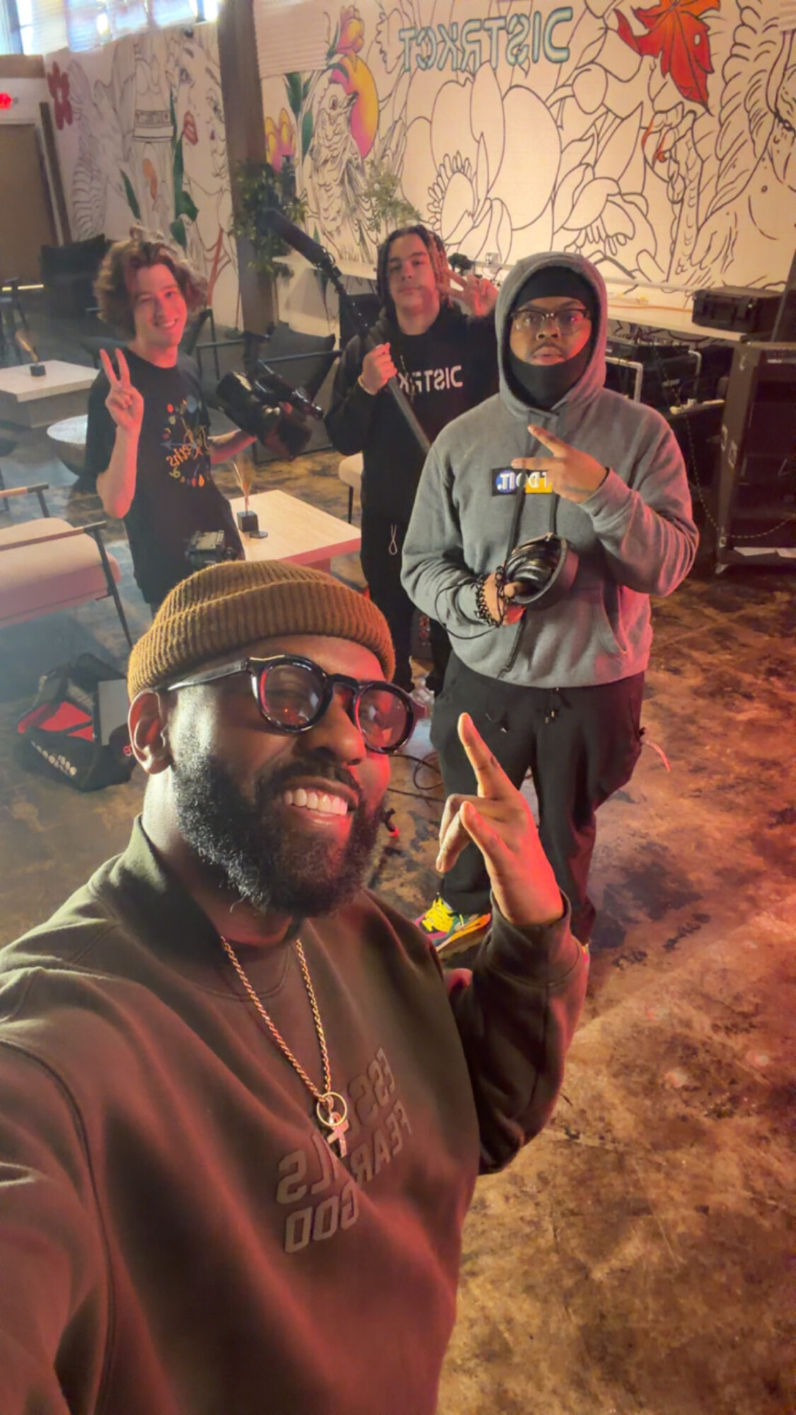 Four people posing indoors, one in front wearing glasses and a hat, three in the background near a mural.