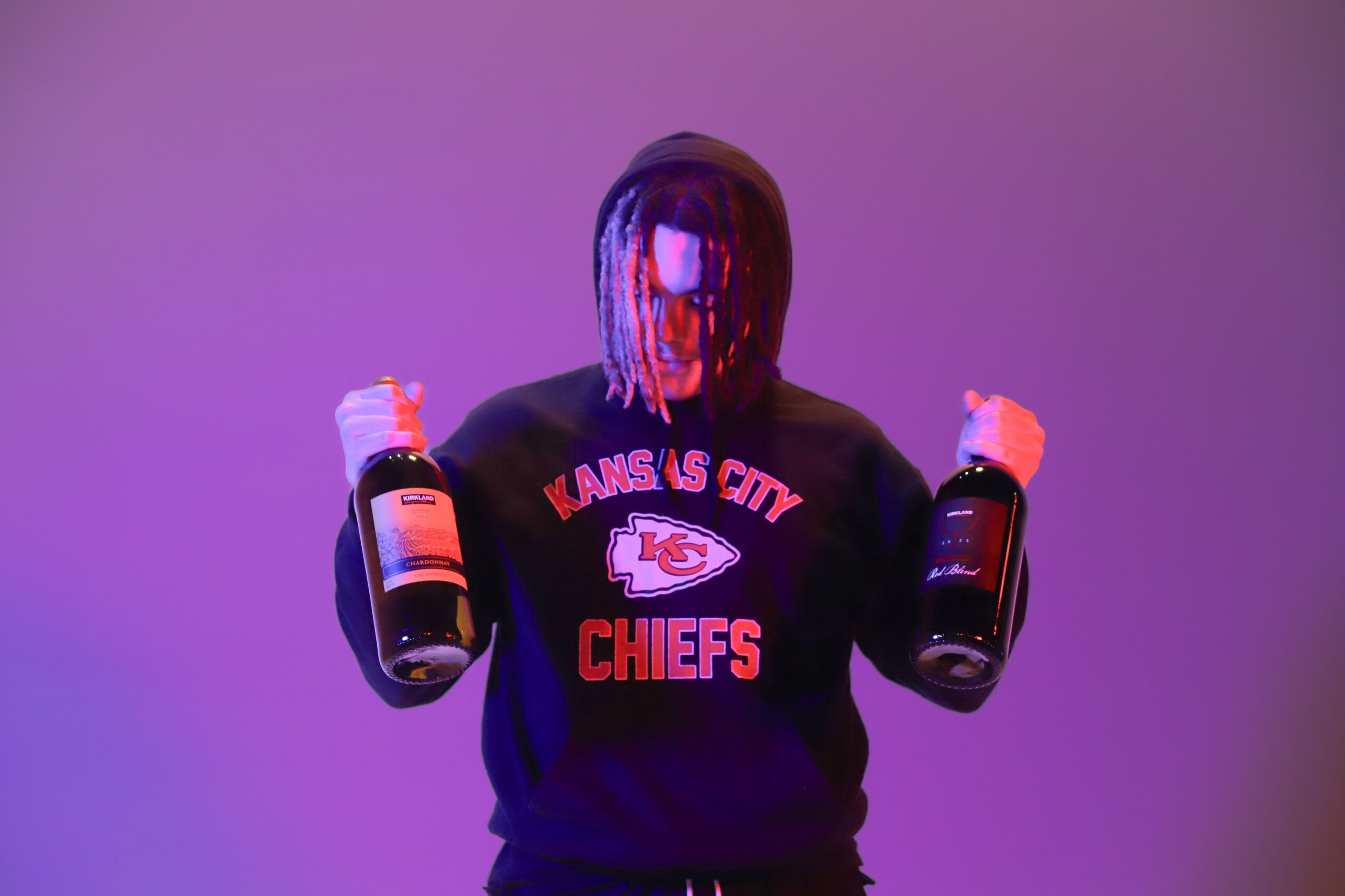Person with dreadlocks holding two bottles, wearing a Kansas City Chiefs sweatshirt, against a purple background.
