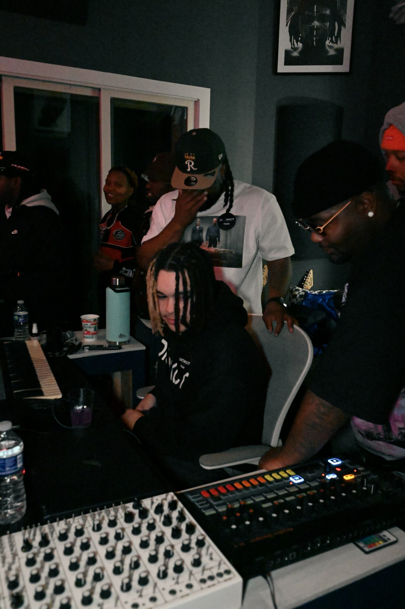 Group of people in a music studio, one person with dreadlocks sitting at a mixing console, others standing around.