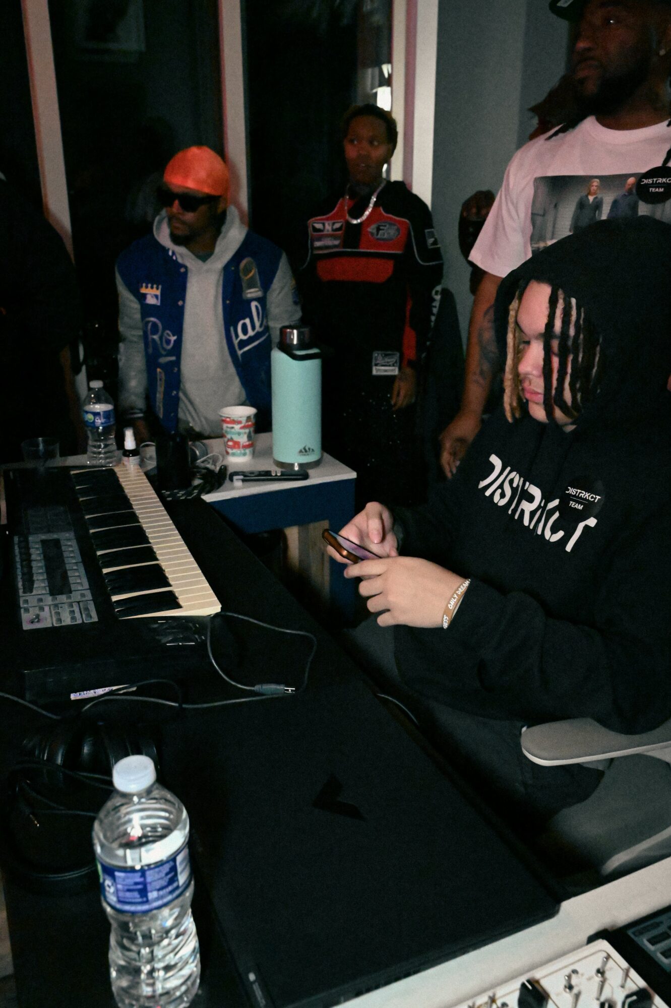 Person with dreadlocks using a laptop at a table with water bottles and electronic equipment, others in the background.