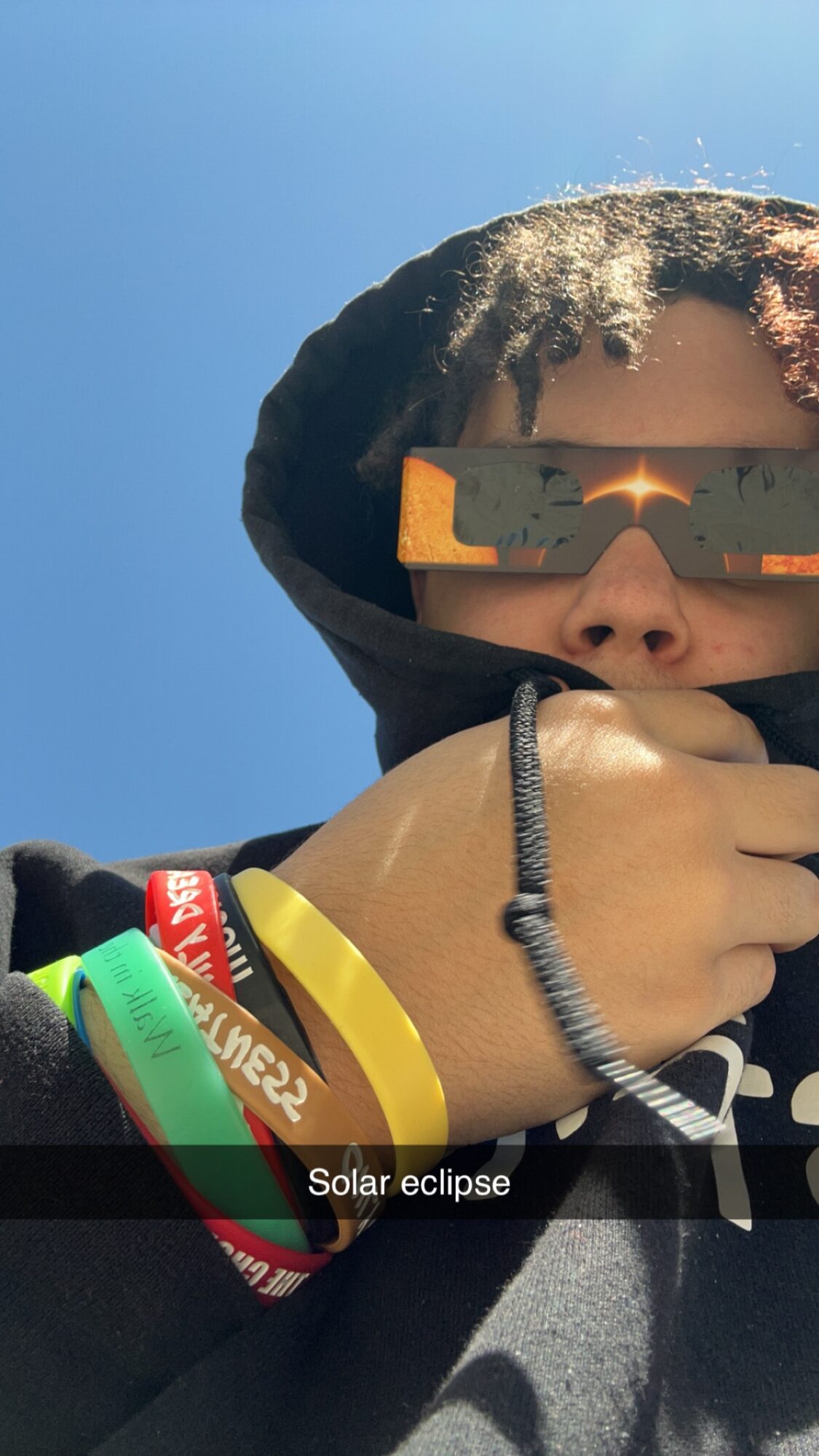 Person wearing sunglasses and a hoodie, holding their chin, with a clear blue sky background.
