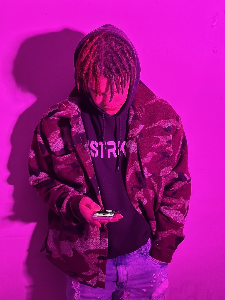 Young person with dreadlocks looking at a phone, wearing a camouflage jacket and hoodie, against a pink background.