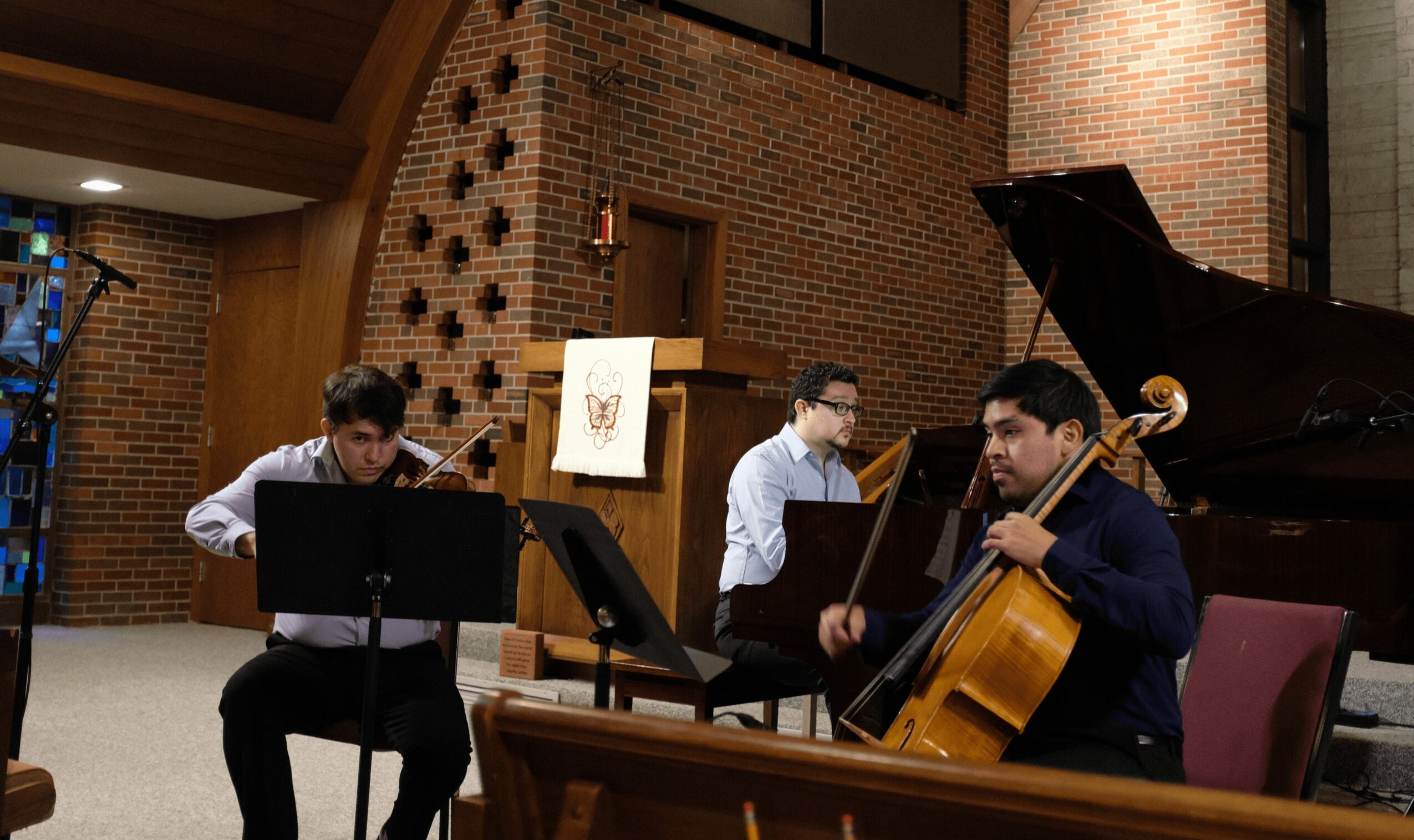 Three musicians perform with a piano, cello, and clarinet in a brick-walled room.