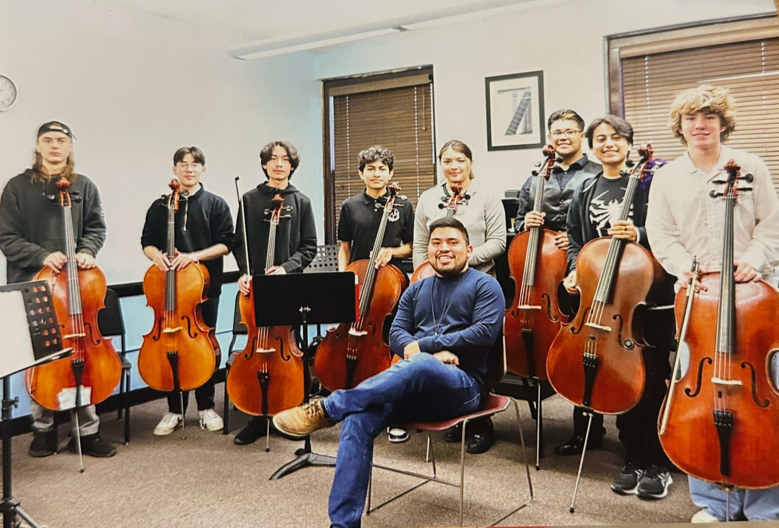 Group of nine people with musical instruments, some holding cellos, in a room with windows and framed pictures.