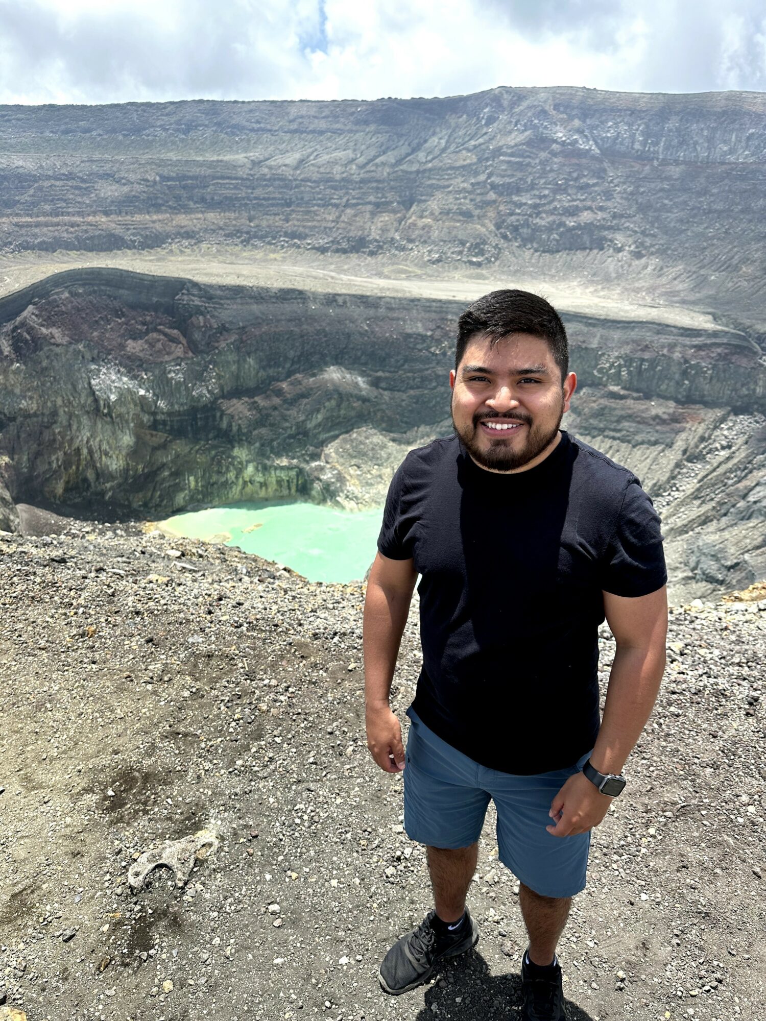 Man standing outdoors with a volcanic crater and green lake in the background.