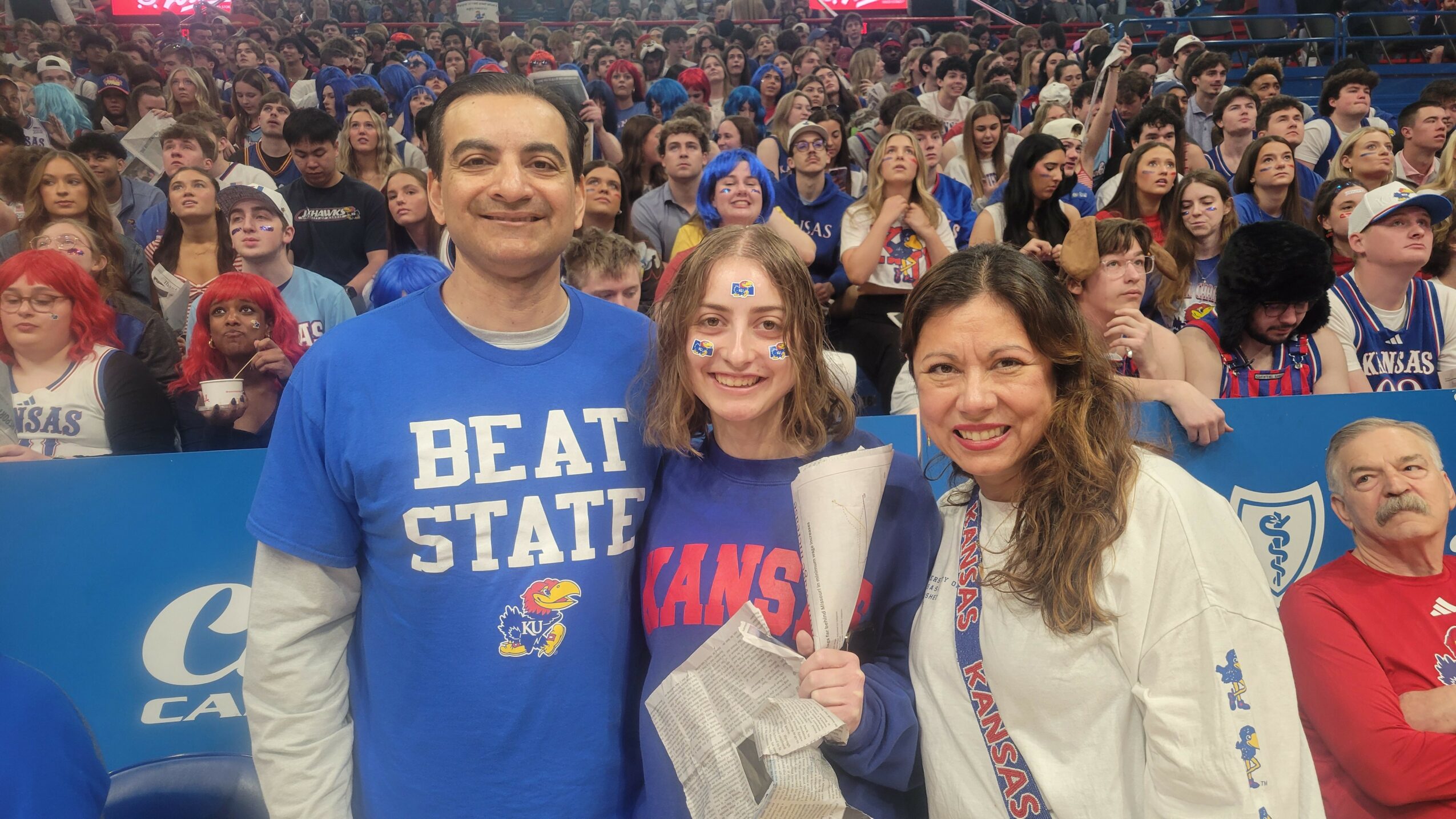 Three people smiling at a crowded event, with many attendees in the background, some wearing blue shirts.