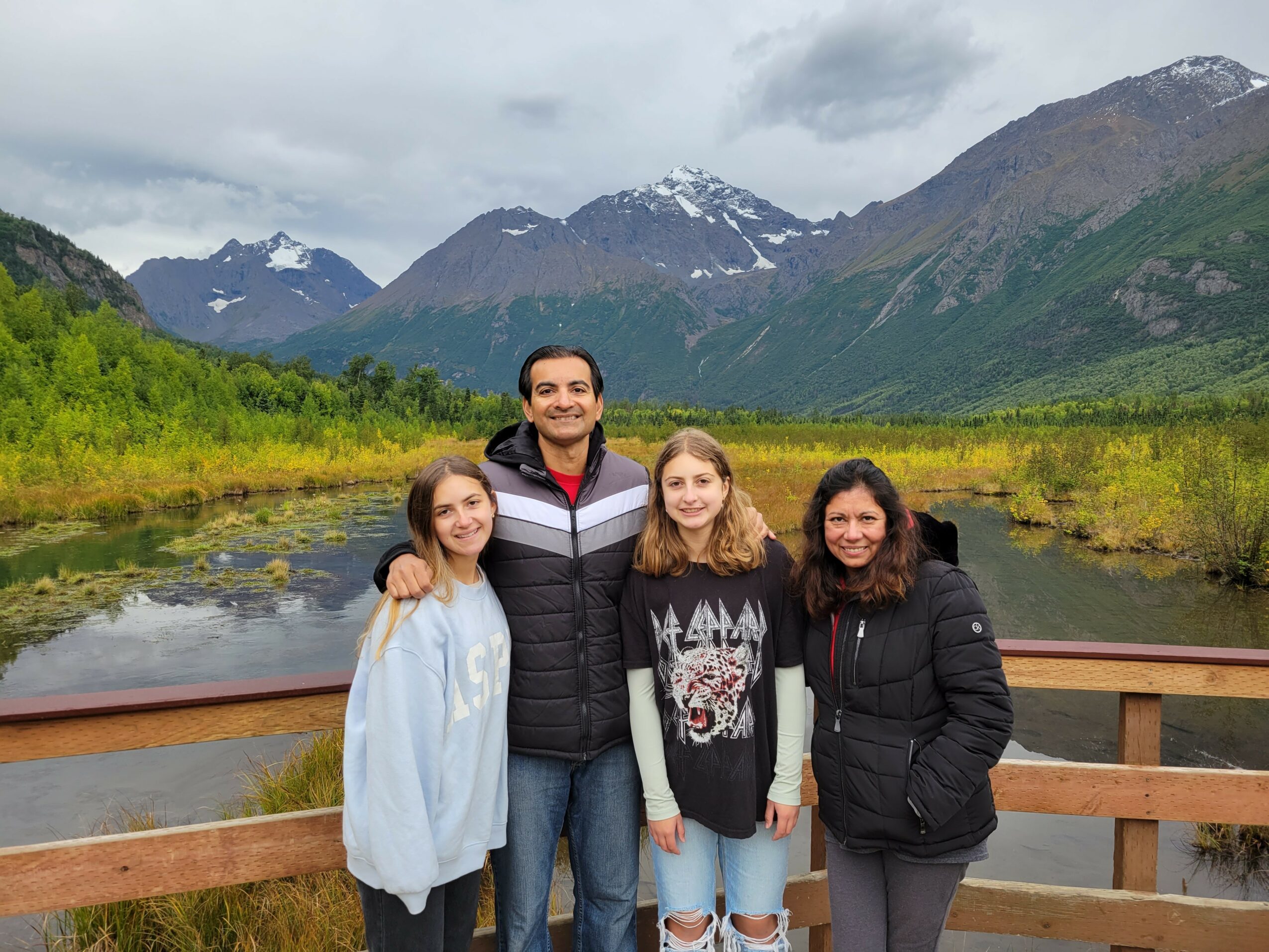 Four people standing on a wooden bridge with mountains and a river in the background, smiling outdoors.