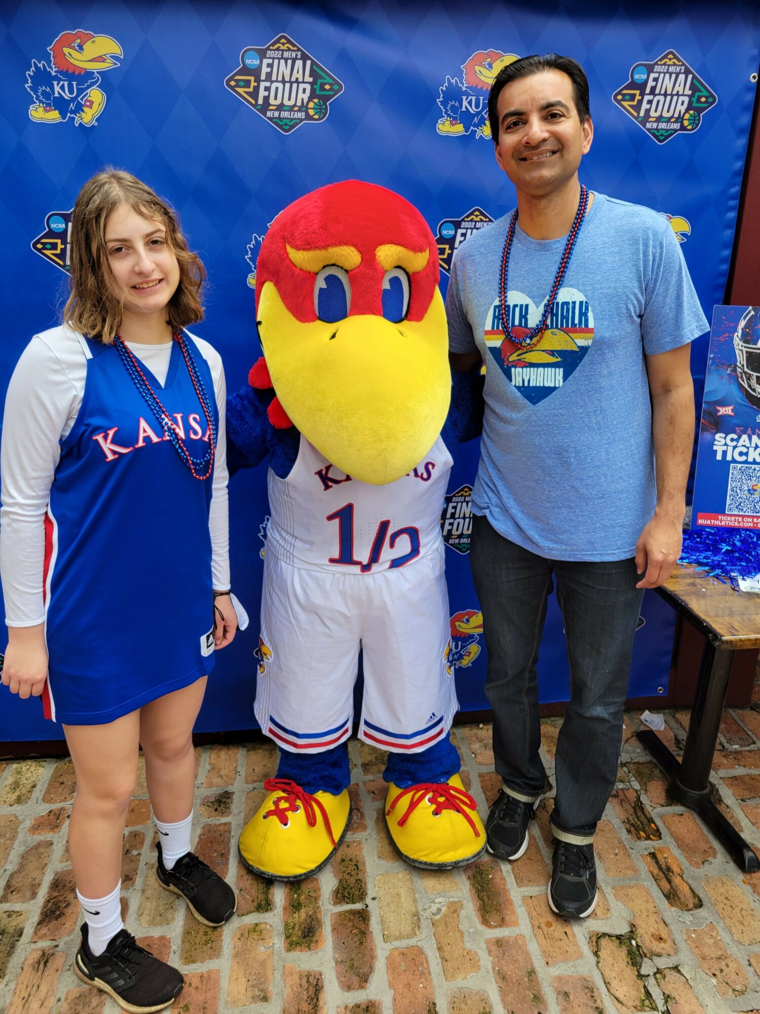 Child in Kansas City Royals jersey with mascot and woman in blue shirt, brick background, group photo.
