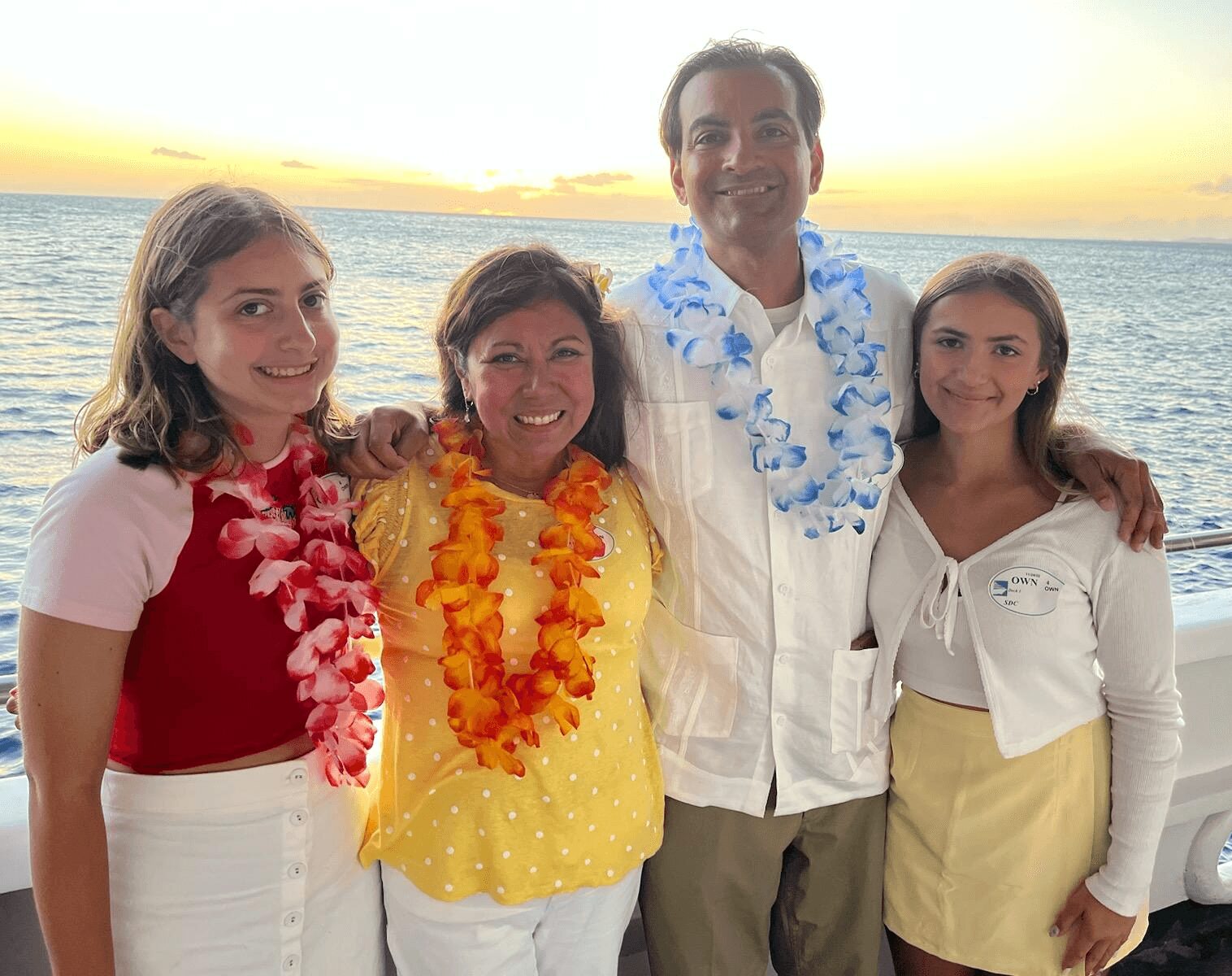 Four people smiling on a boat at sunset, wearing leis and casual clothing, with ocean in background.