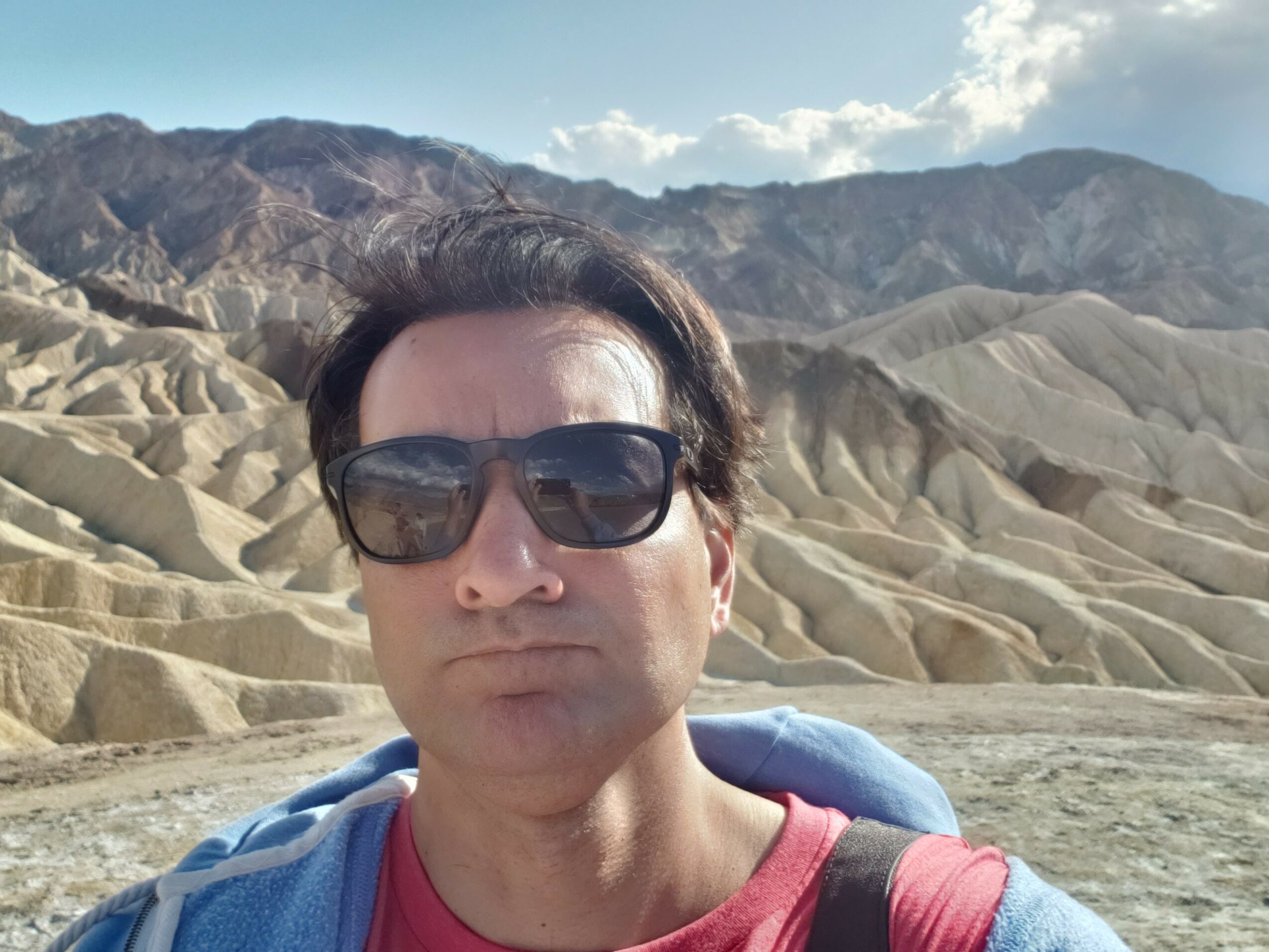 Person wearing sunglasses and a red shirt outdoors with mountainous terrain in the background.