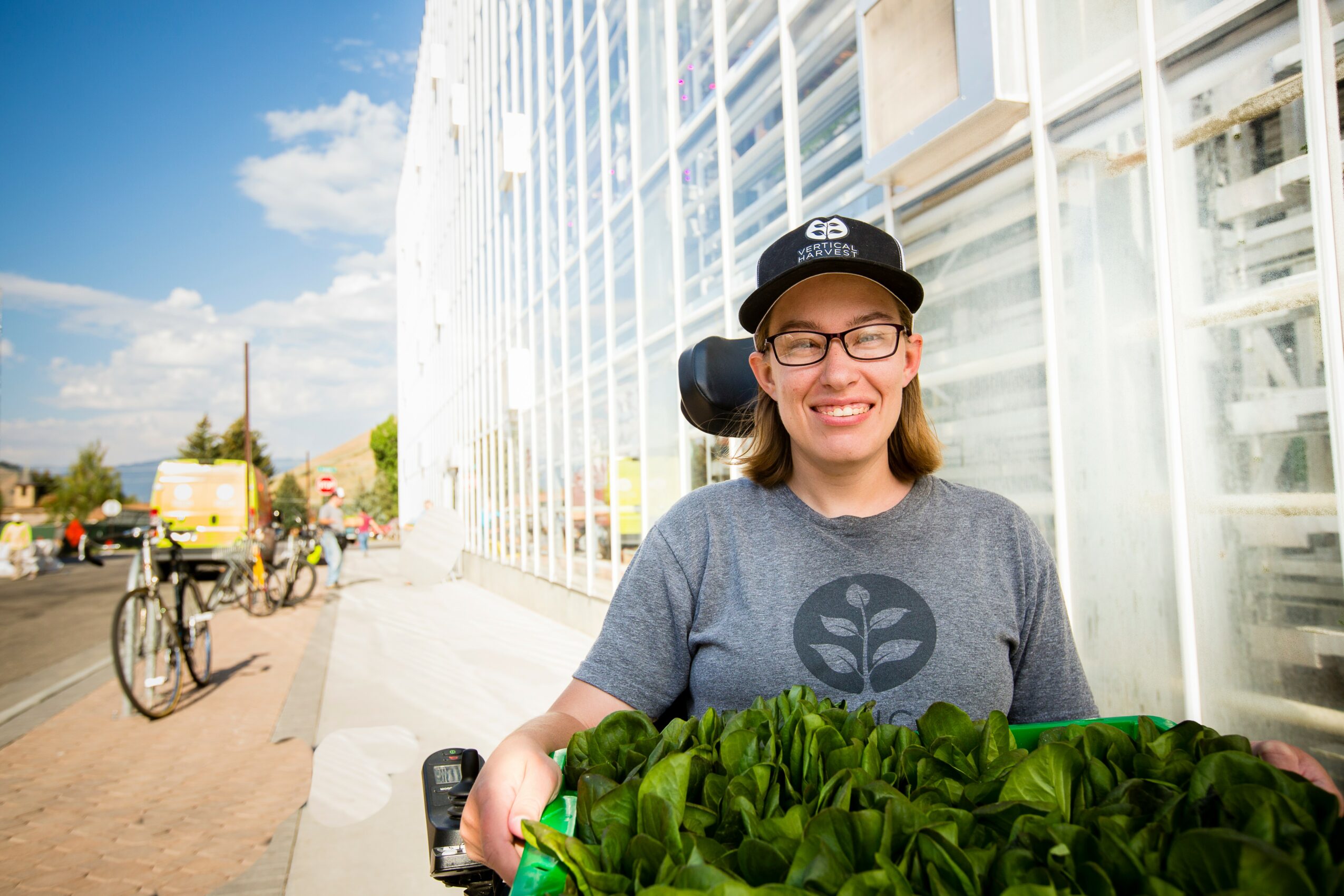 Smiling woman with glasses and a cap standing outside near a glass building, holding a basket of leafy greens.