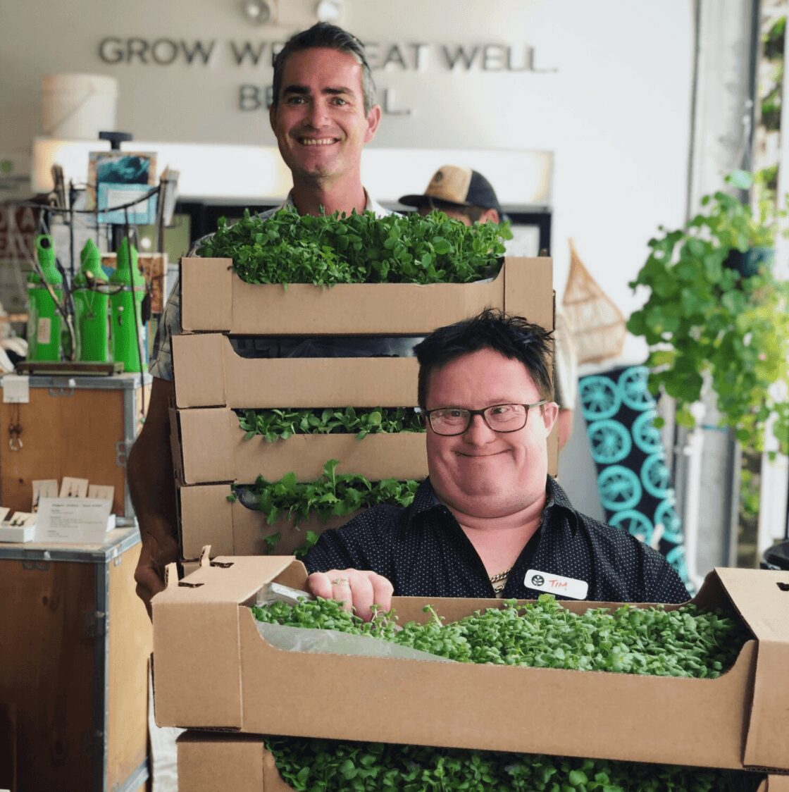 Two men with boxes of green plants inside a store, smiling at the camera, with shelves and plants in the background.