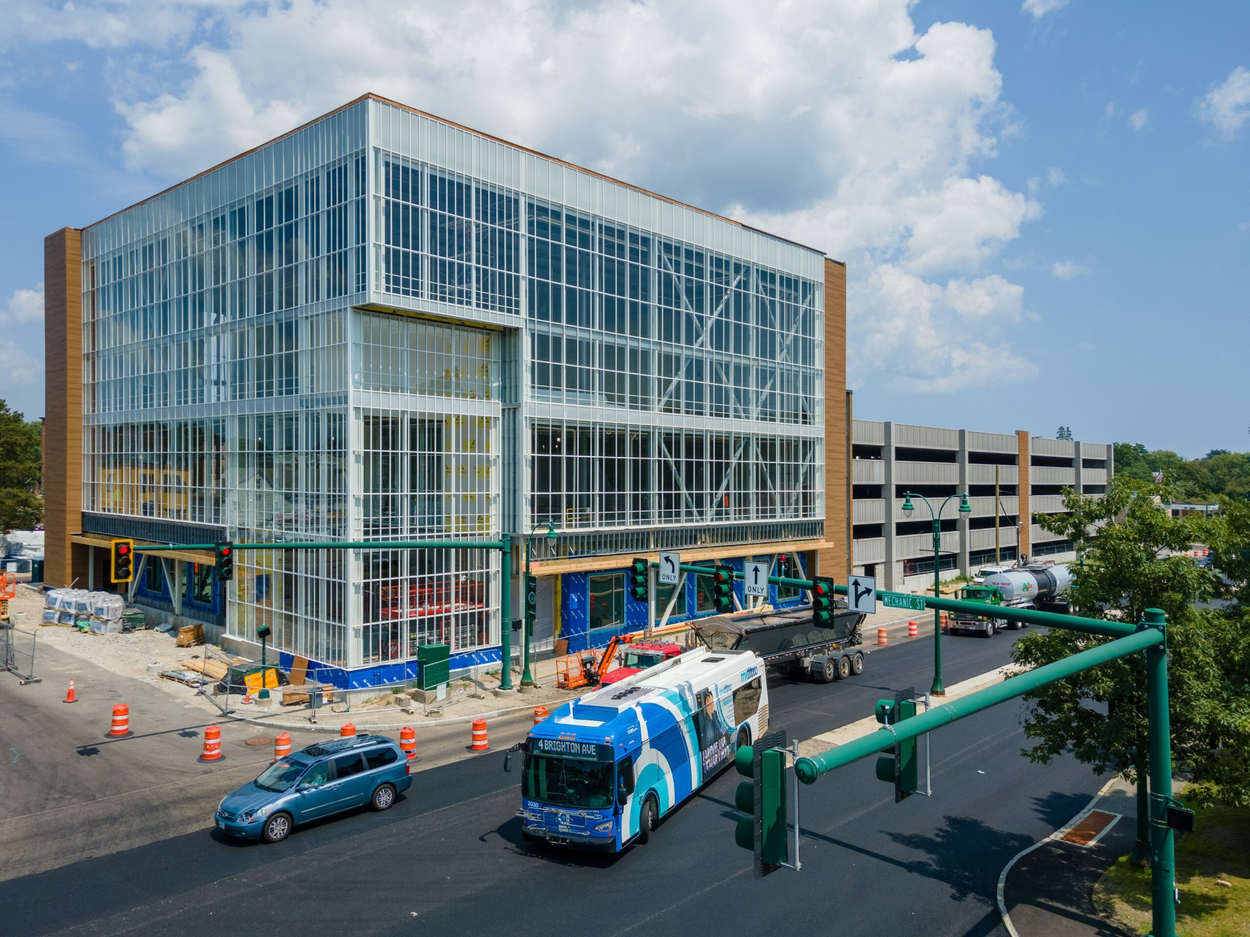 Modern glass building with a parking garage nearby, vehicles on the street, and a blue sky with clouds.