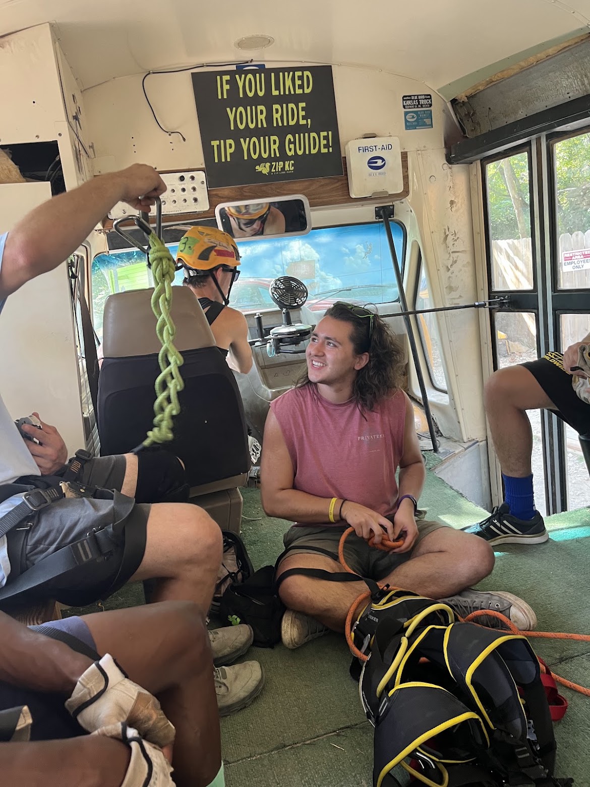 Group of people sitting inside a bus, with a woman in a pink shirt smiling and holding a helmet, others around her.