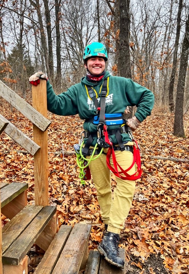 Person in outdoor gear standing on leaf-covered ground, smiling, with trees in background, wearing a helmet and harness.
