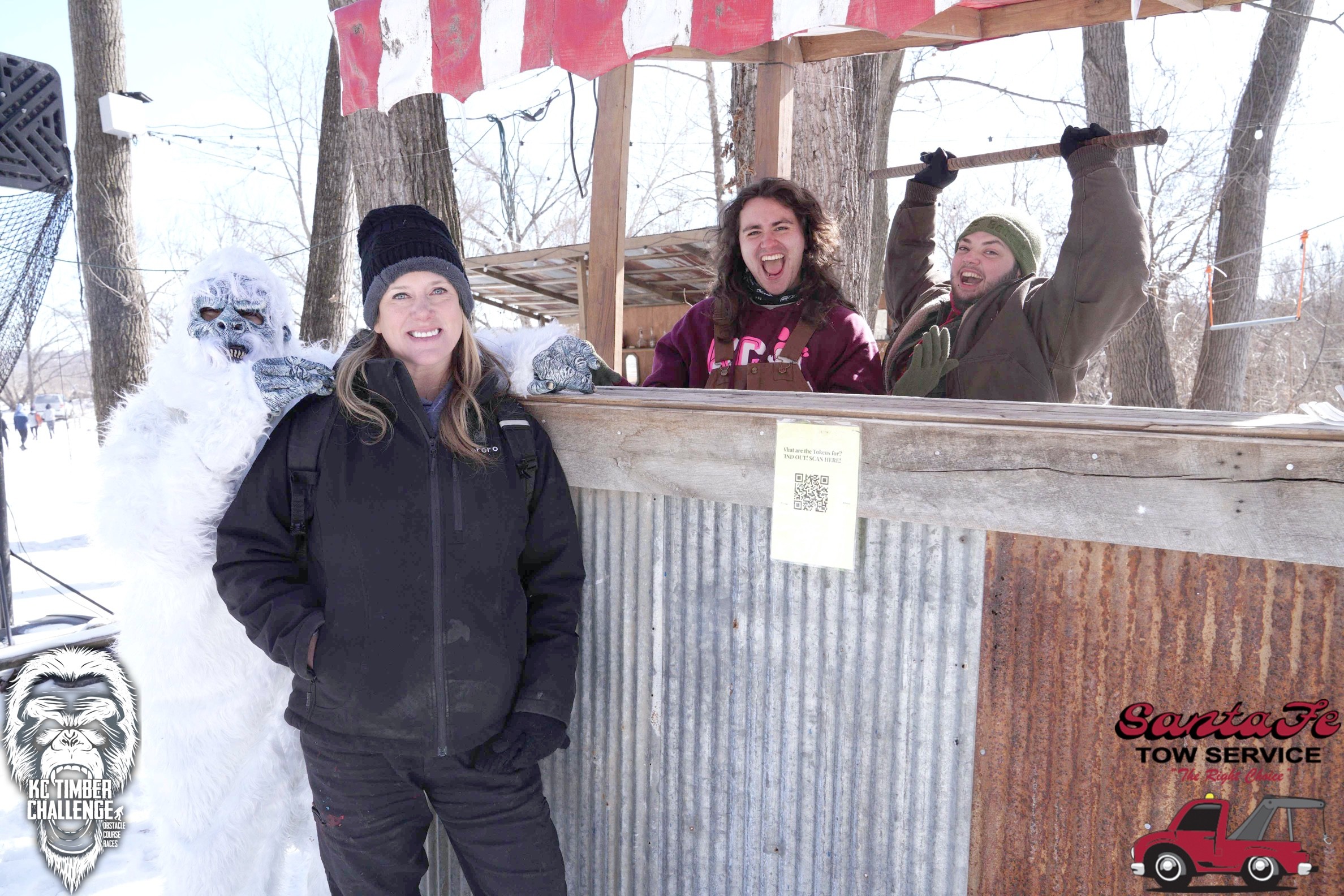 Three people standing behind a wooden and metal structure outdoors in winter, with snow and trees in the background.