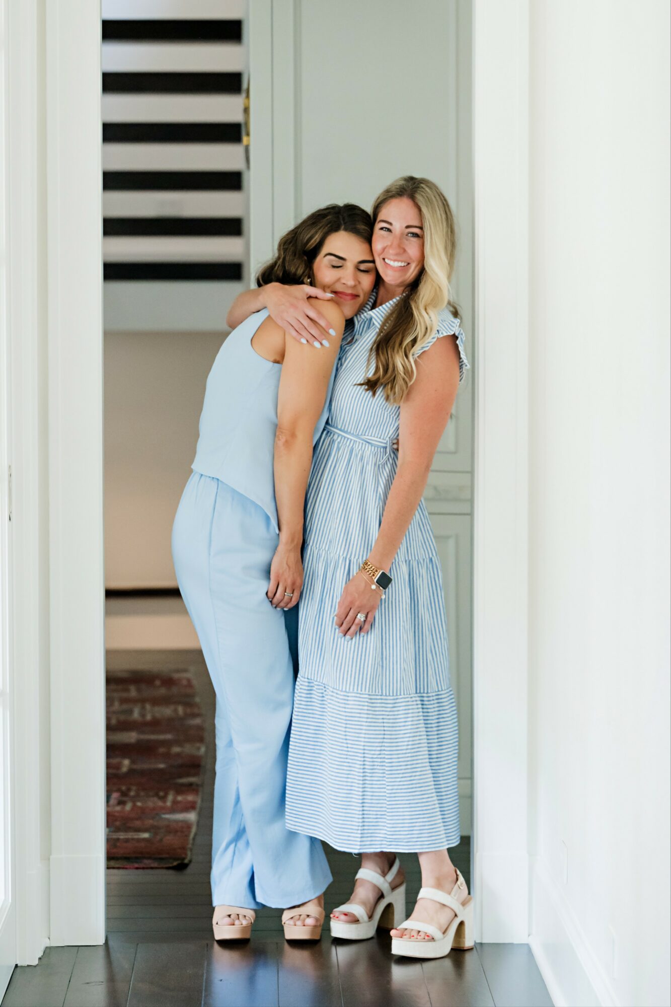 Two women hugging and smiling in a doorway, one in a light blue jumpsuit, the other in a blue dress, both wearing heels.