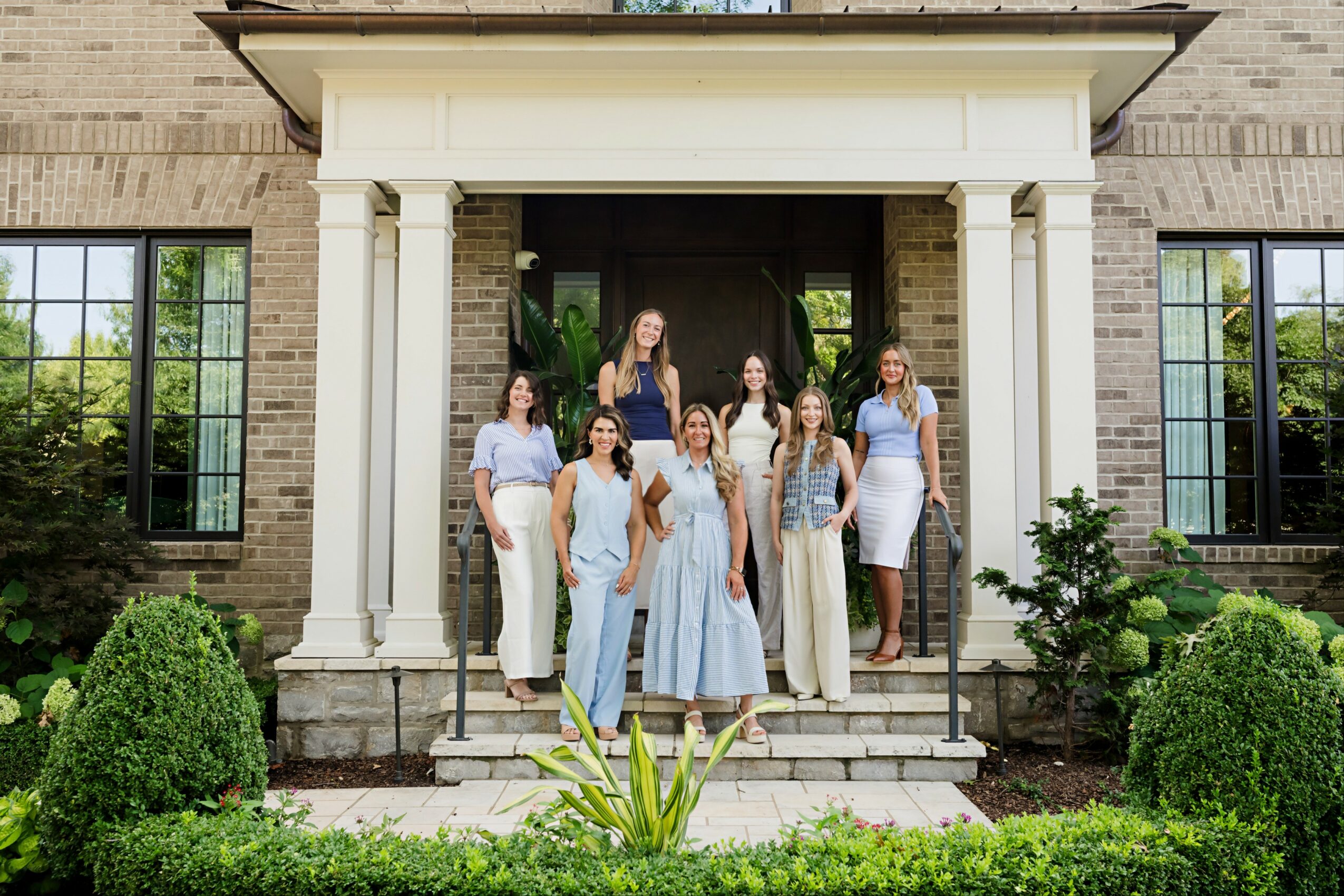 Seven women standing on steps in front of a building entrance, surrounded by greenery and bushes.