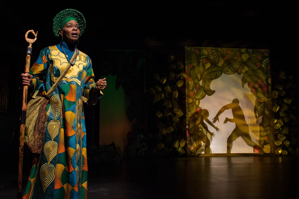 Performer in colorful costume on stage with shadow puppets in background, holding staff, expressive pose.