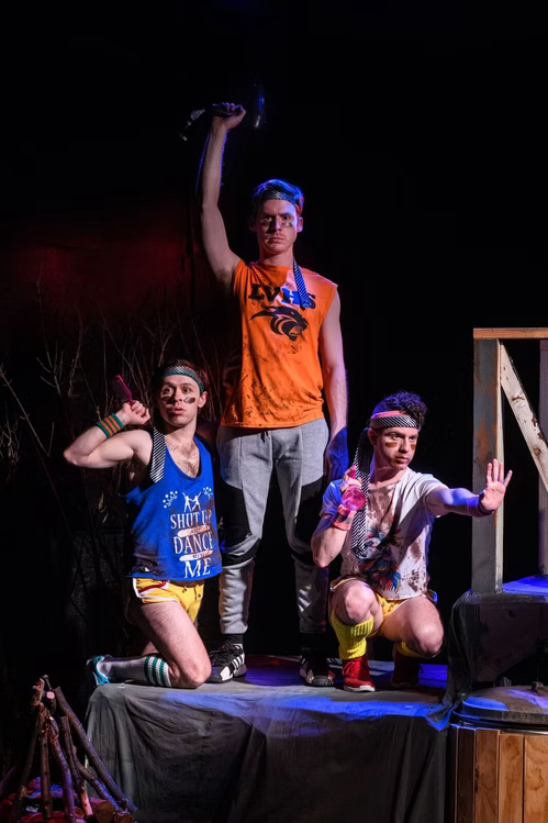 Three children on stage, one standing with arm raised, two kneeling, in colorful costumes, with a dark background.