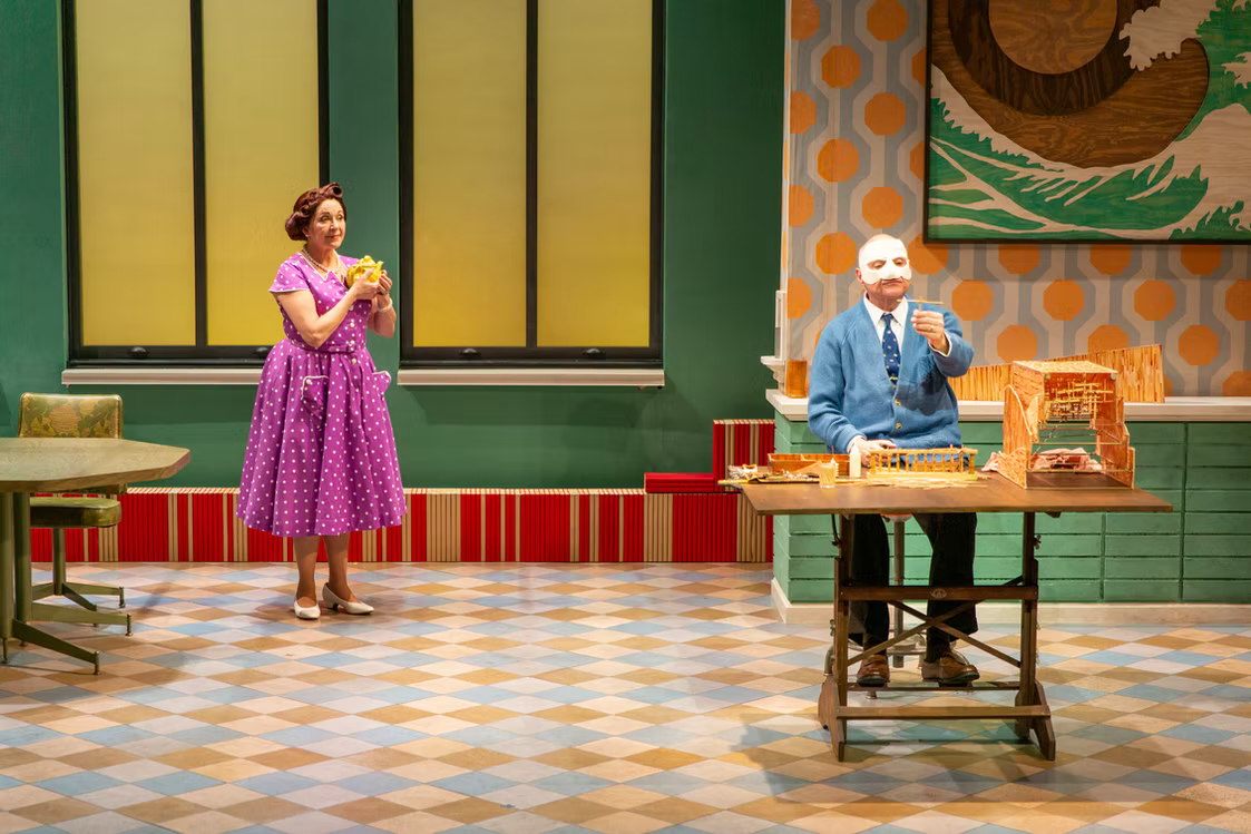 Woman in purple dress and man in blue suit on stage with colorful background and patterned floor.