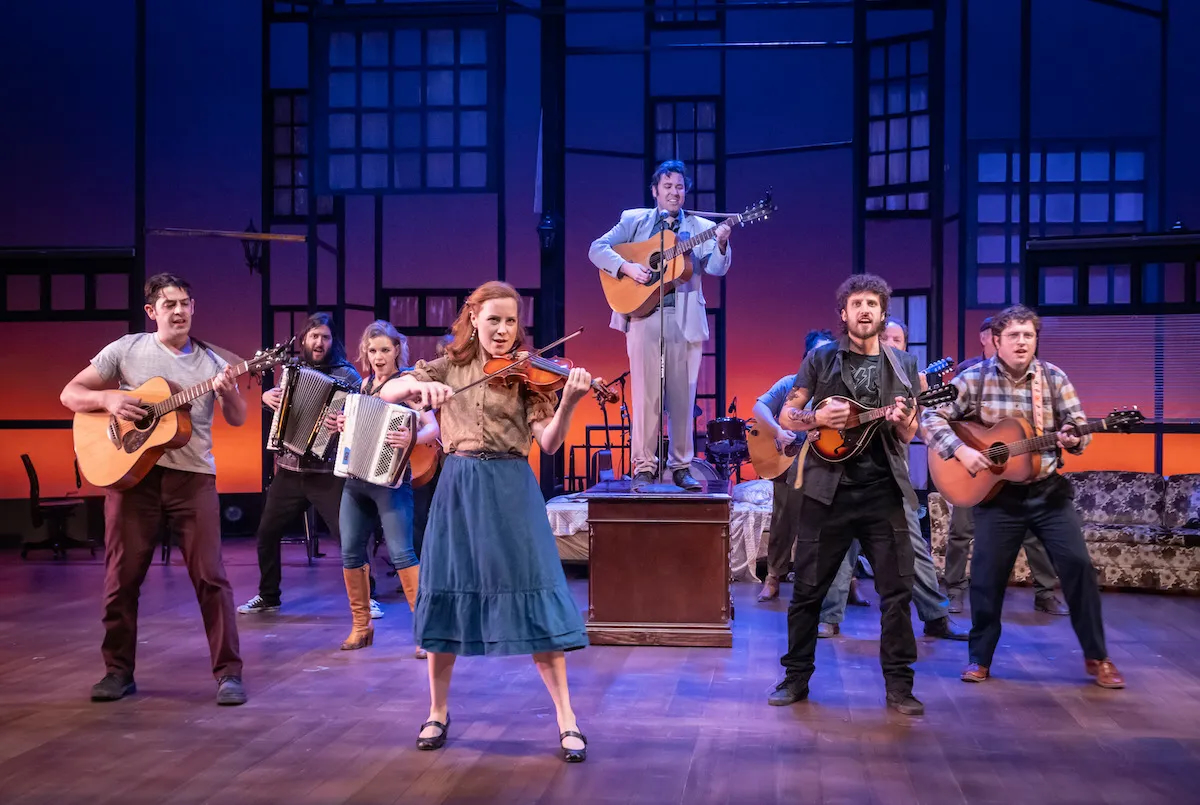 Group of seven performers on stage, playing guitars, accordion, and singing, with a blue-lit background and stage set.