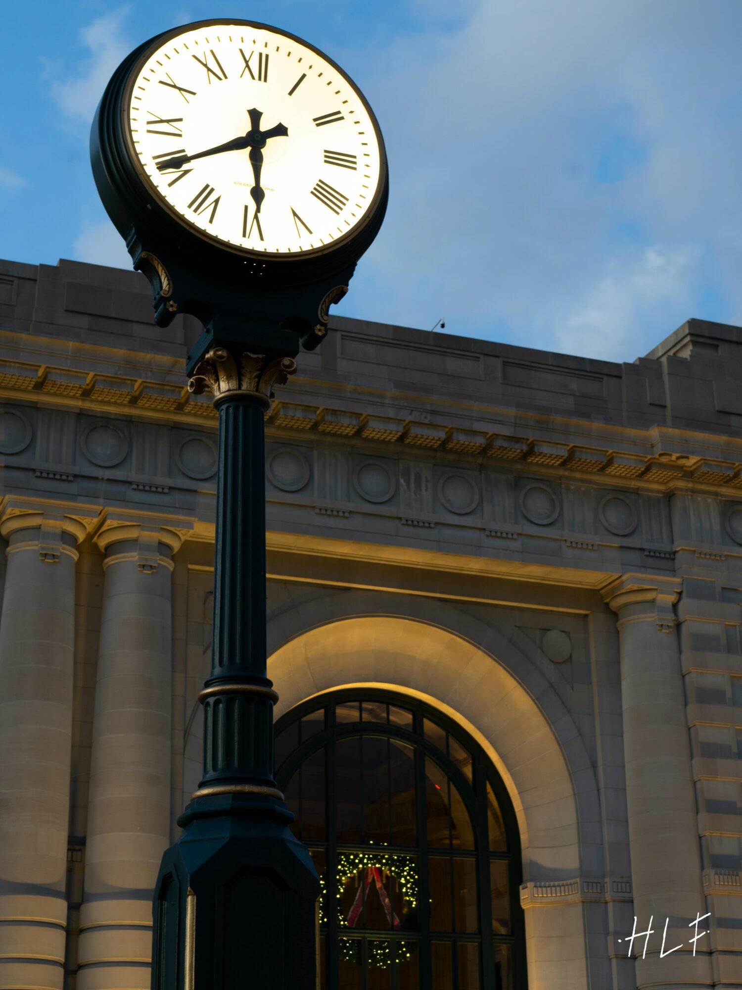 Clock on a tall post shows 5:30, building with arched window and columns in background, blue sky.