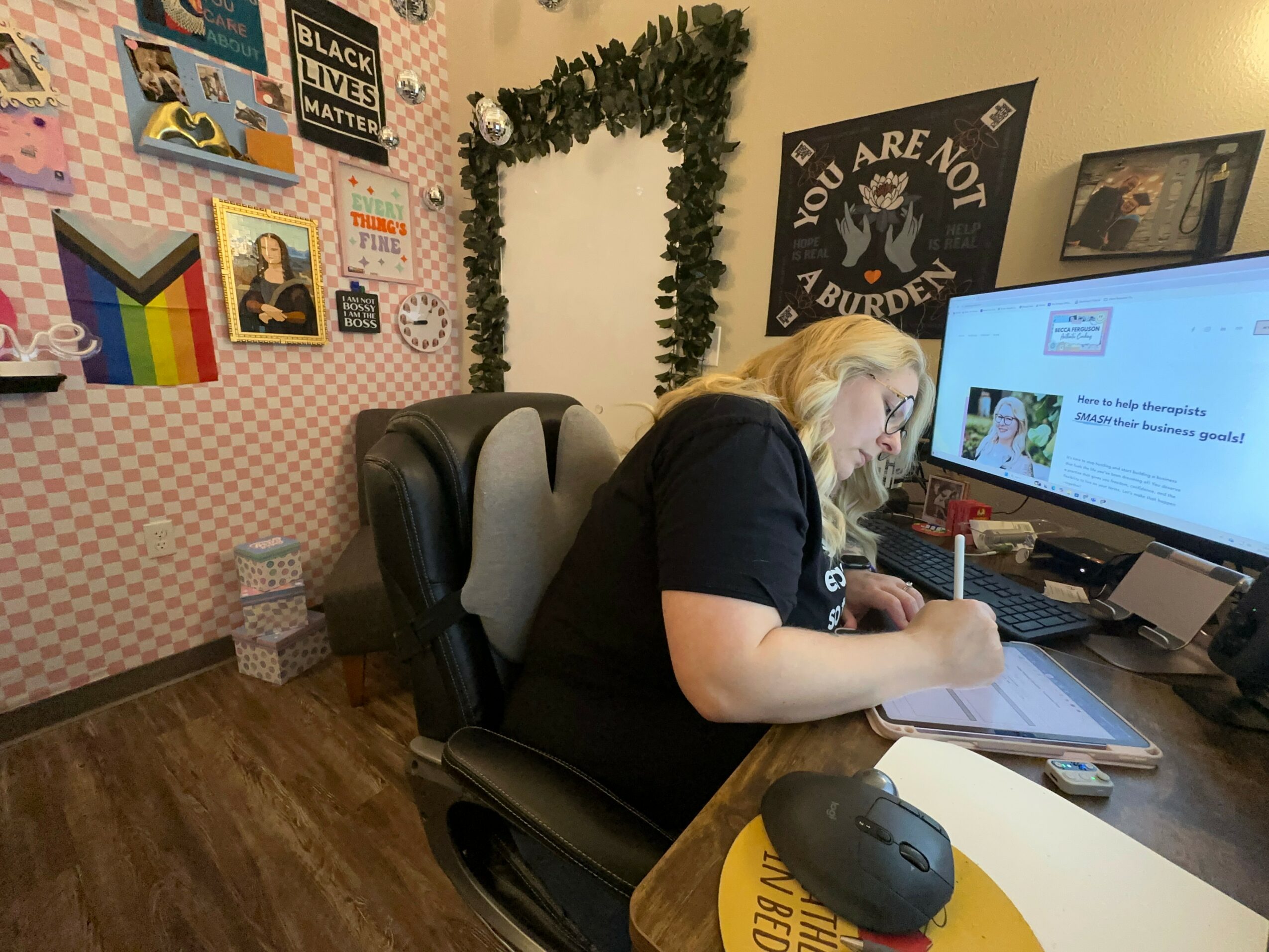 Person with blonde hair working at a desk with a computer, surrounded by posters and decorations on the wall.