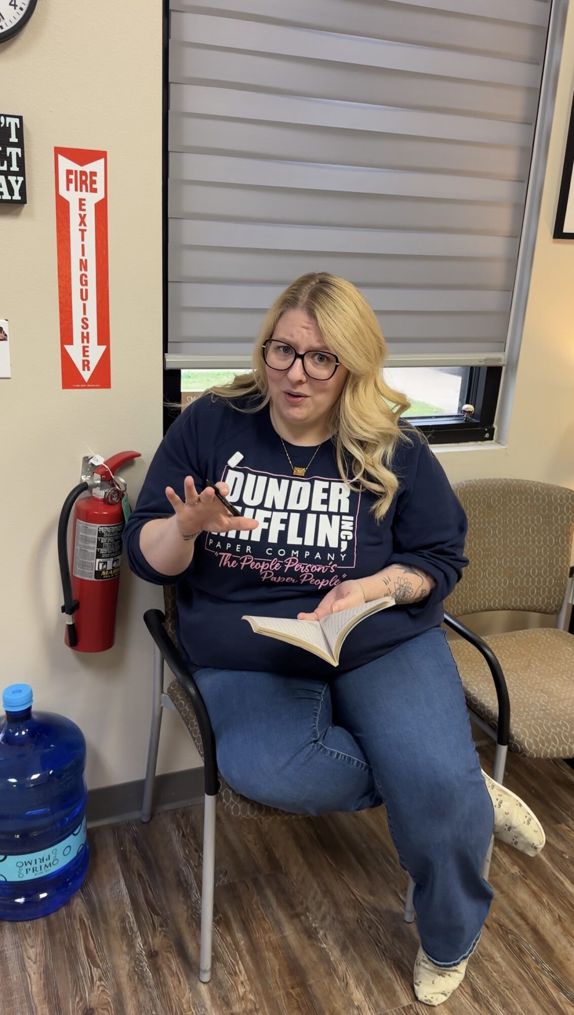 Woman with glasses and blonde hair sitting in a chair, holding a book and making a peace sign, in a room with a window and fire extinguisher.