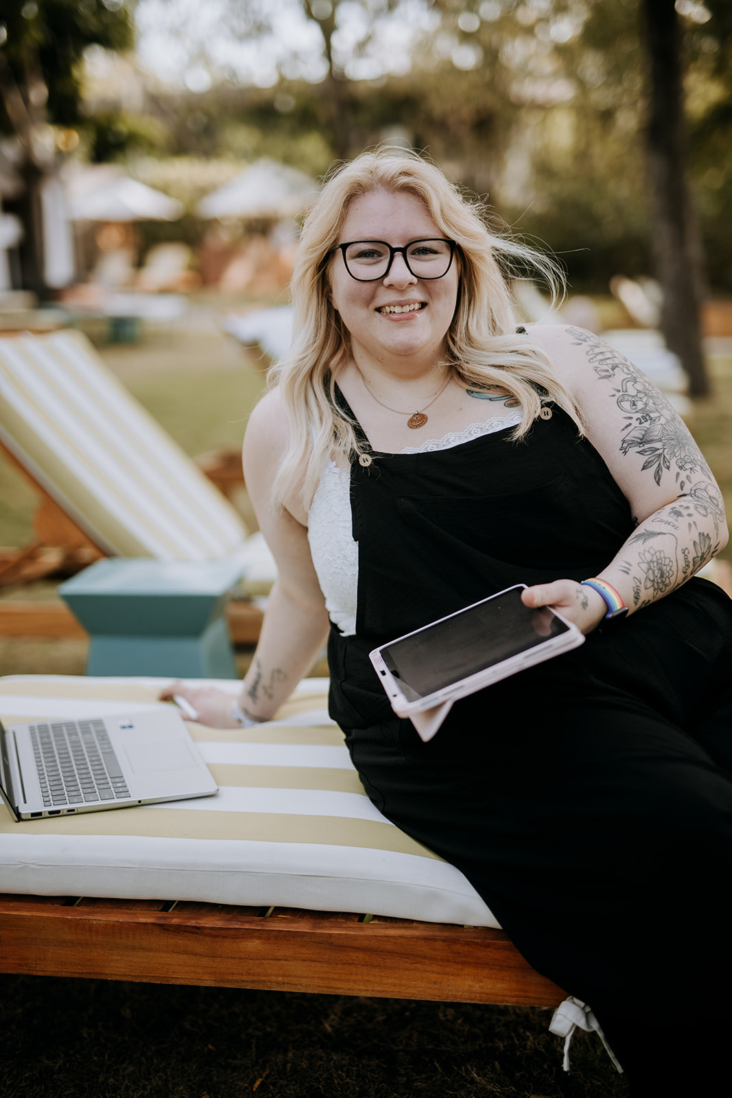 Woman with glasses and long blonde hair sitting outdoors on a lounge chair, holding a tablet, smiling, with a laptop nearby.