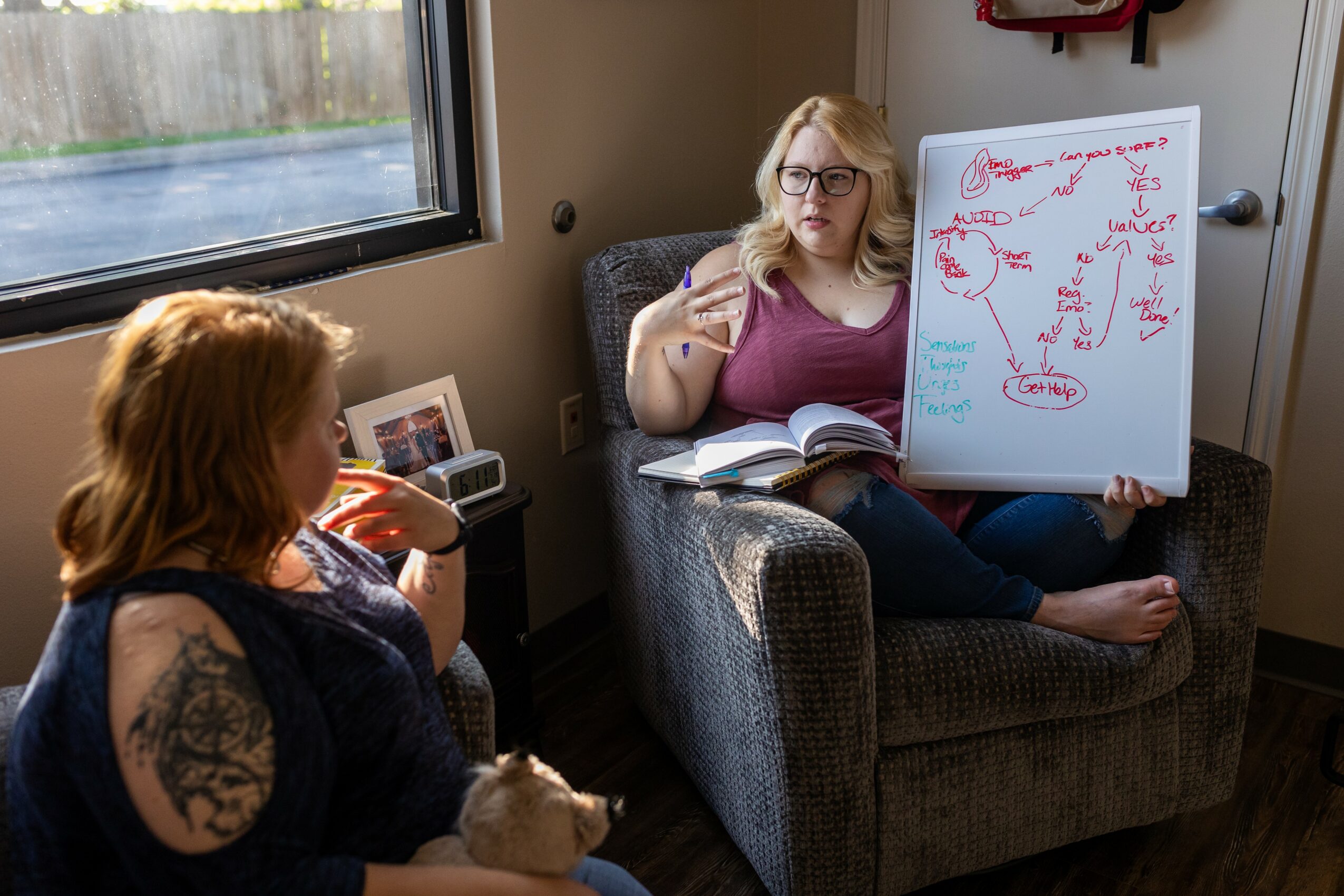 Two women in a room, one sitting in a chair with a whiteboard, the other drinking from a glass, window in background.