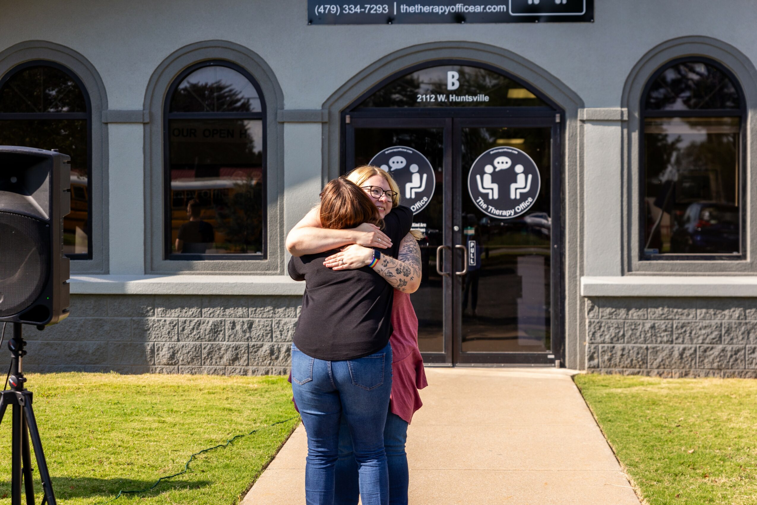 Two people hugging outside a building with arched windows and a door with seating icons, on a sidewalk with grass.