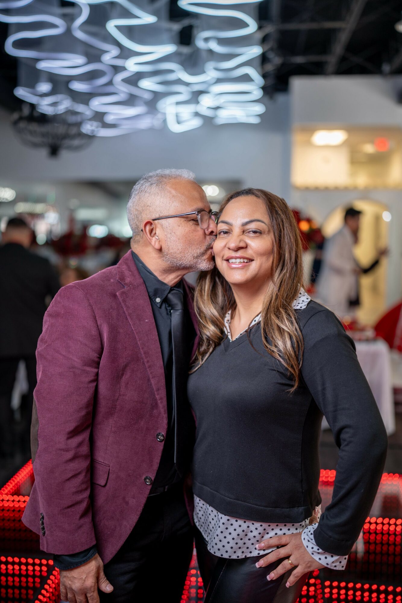 Older man kissing young woman on the cheek at an indoor event, smiling, with decorative lighting overhead.