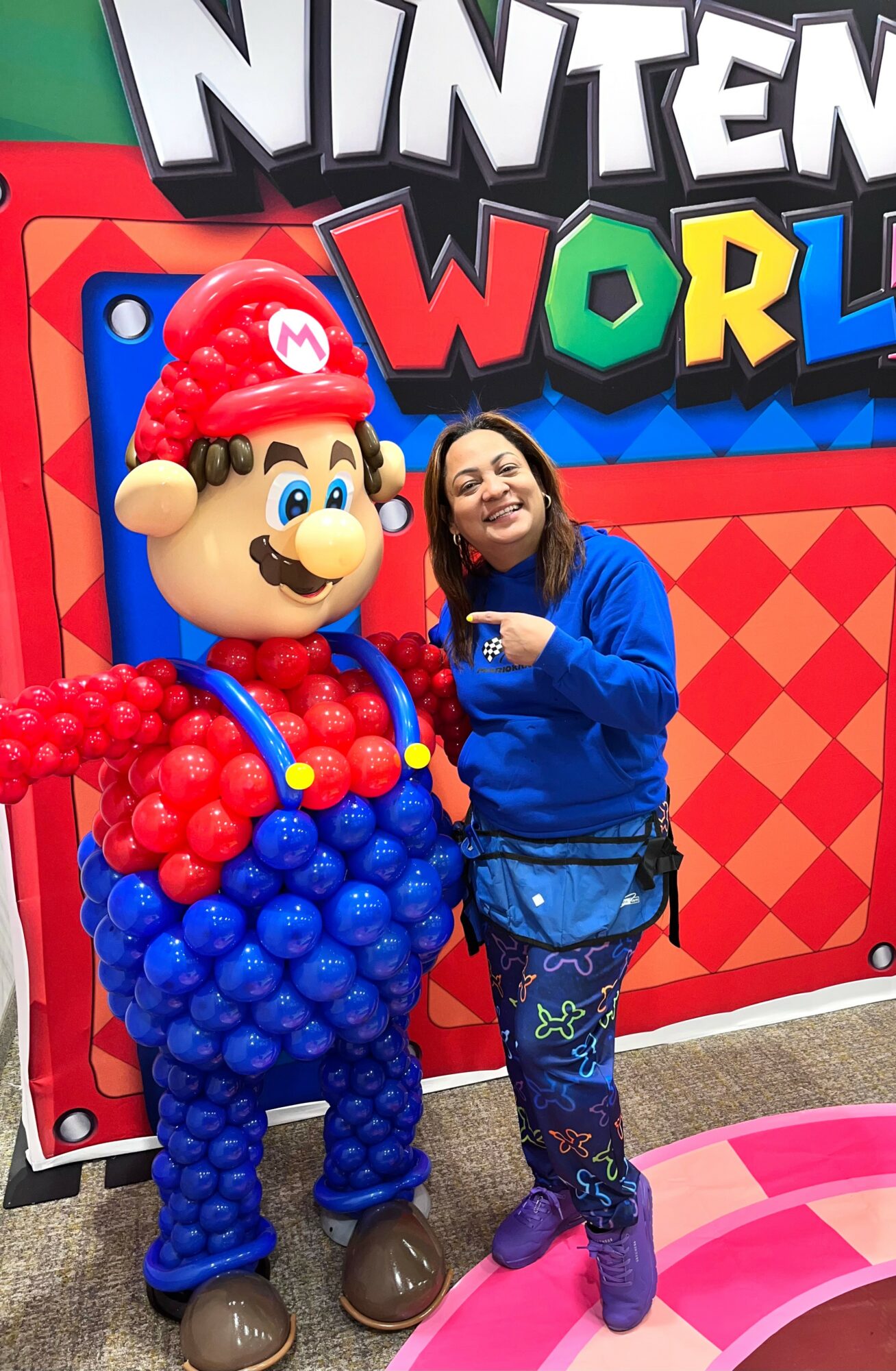 Person in blue jacket and patterned pants standing next to a Mario balloon figure at Nintendo World, colorful background.