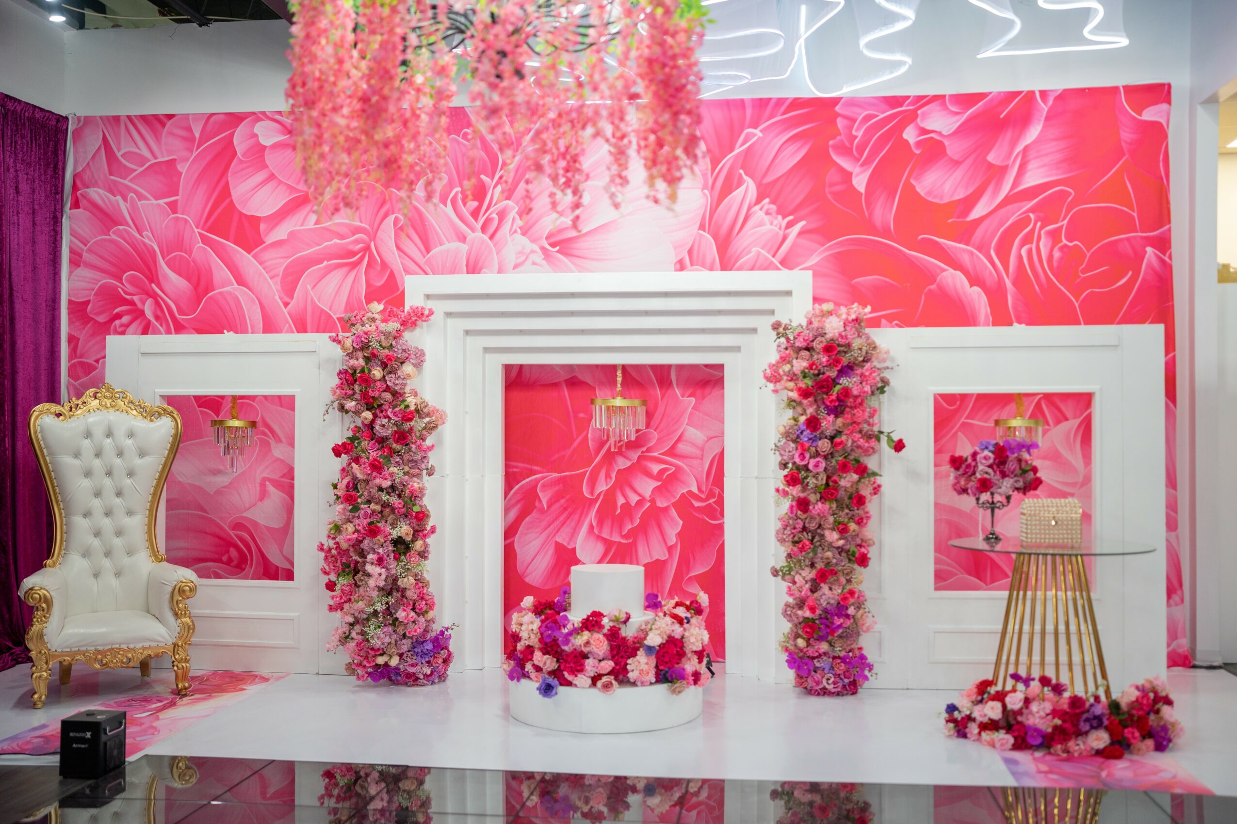 Decorative pink floral backdrop with white frame, floral arrangements, a white chair, and a small table with flowers.