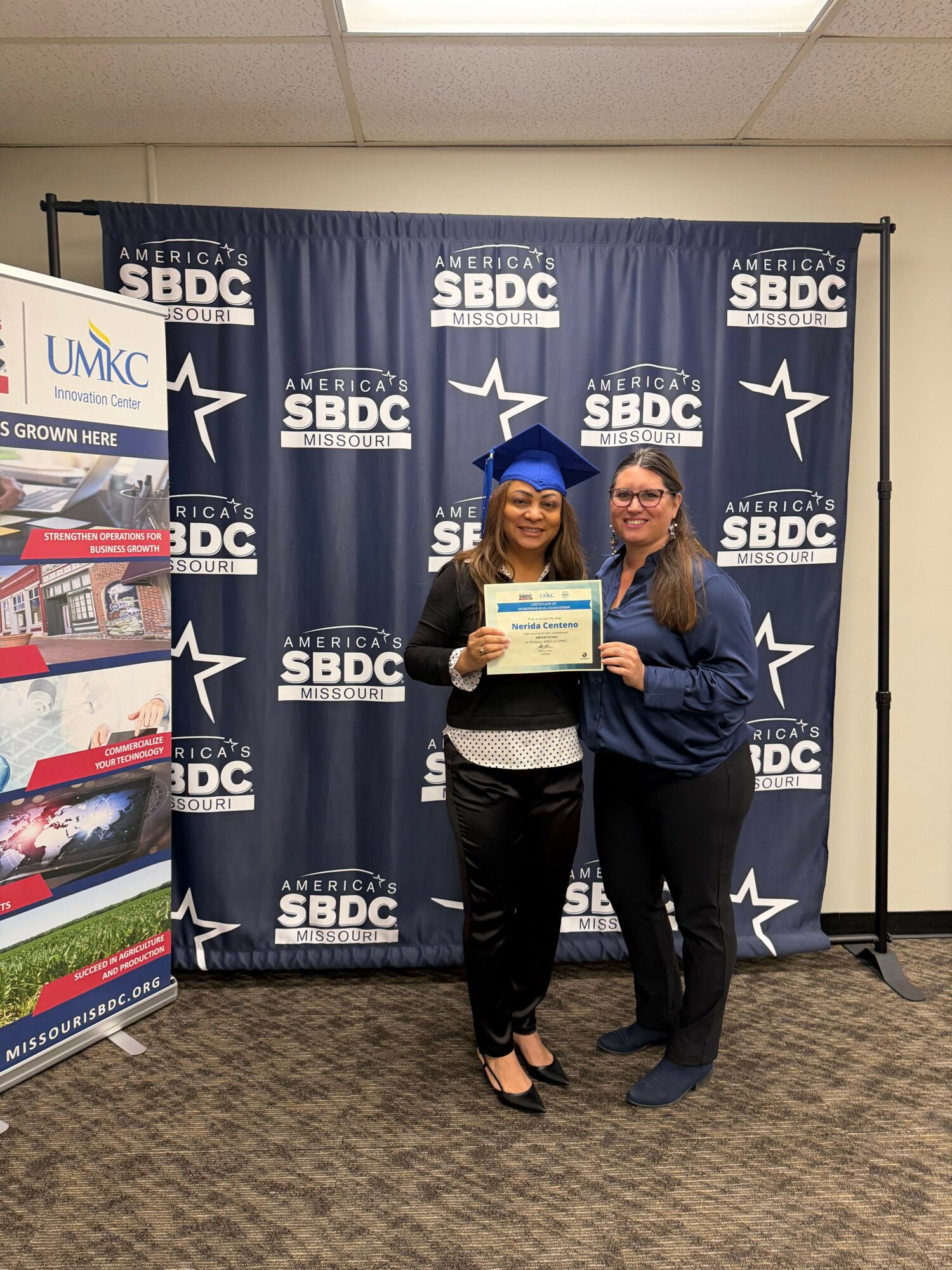 Two women standing in front of a blue SBIDC backdrop, one holding a certificate, both smiling, in a professional setting.
